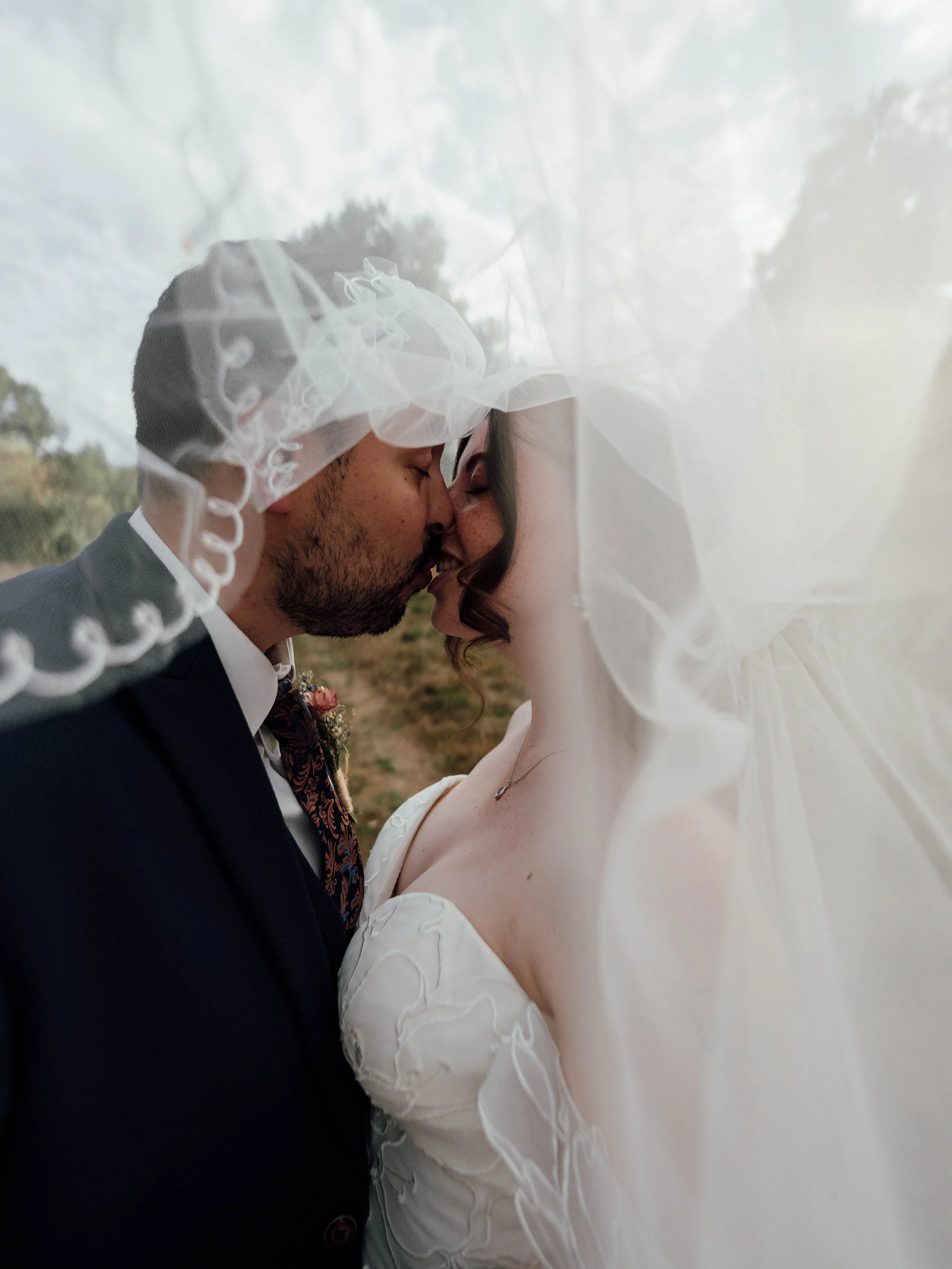 A close-up of a wedding couple sharing a kiss, with the bride's veil covering both of their faces, outdoors with trees and sky in the background.