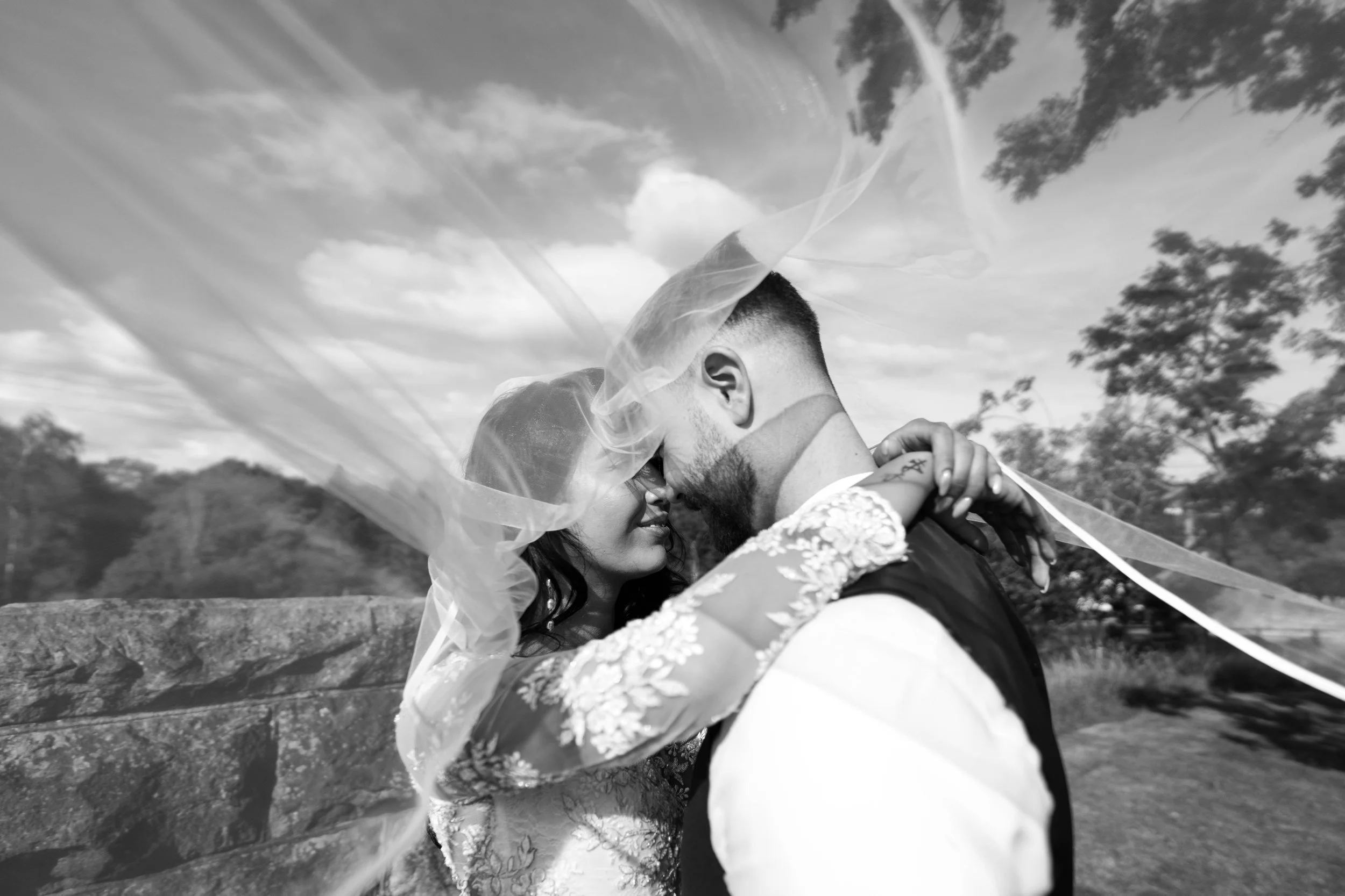 Black and white photo of a bride and groom embracing closely with foreheads touching under a veil outdoors, with trees and cloudy sky in the background.