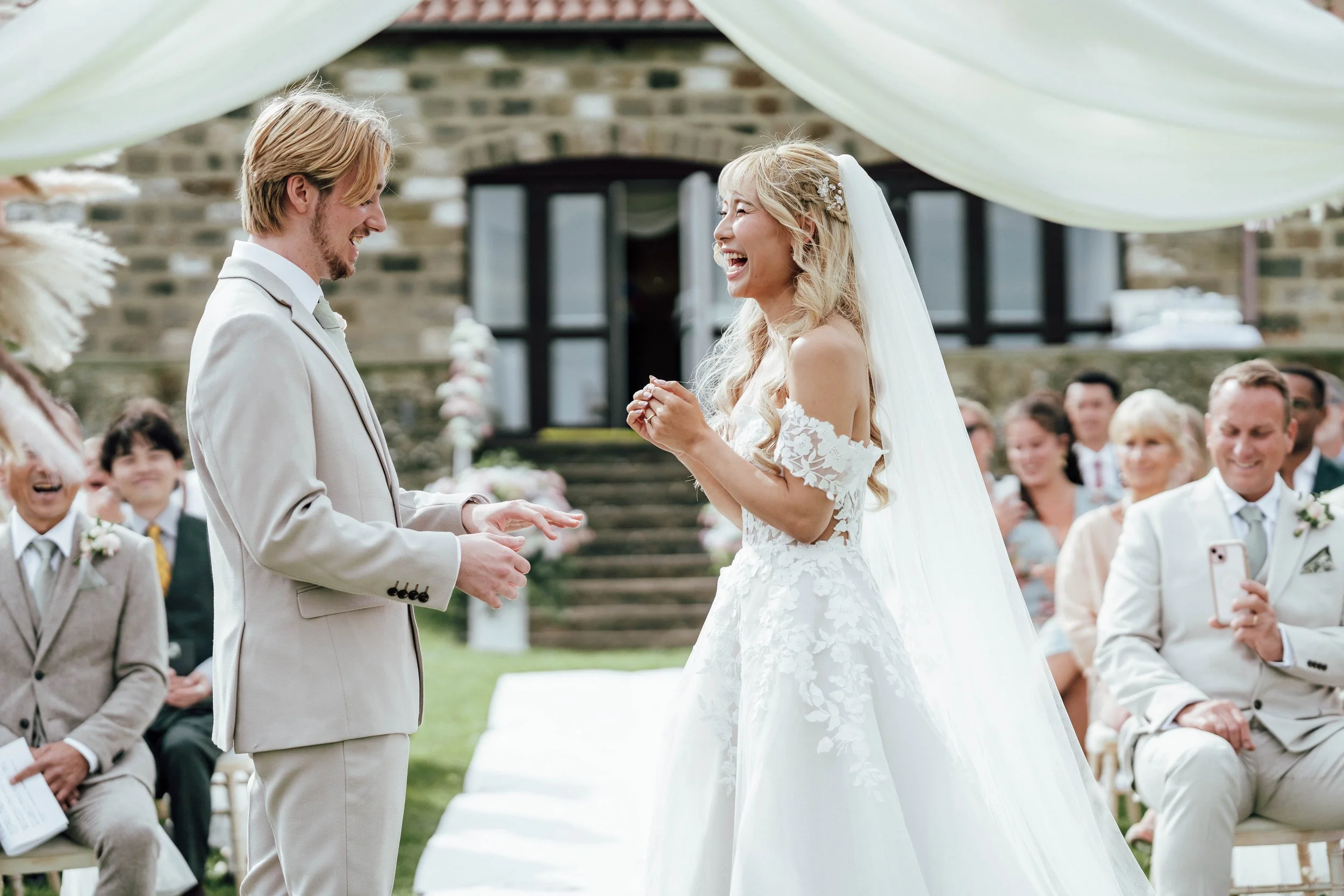 Bride and groom smiling during outdoor wedding ceremony, surrounded by seated guests.