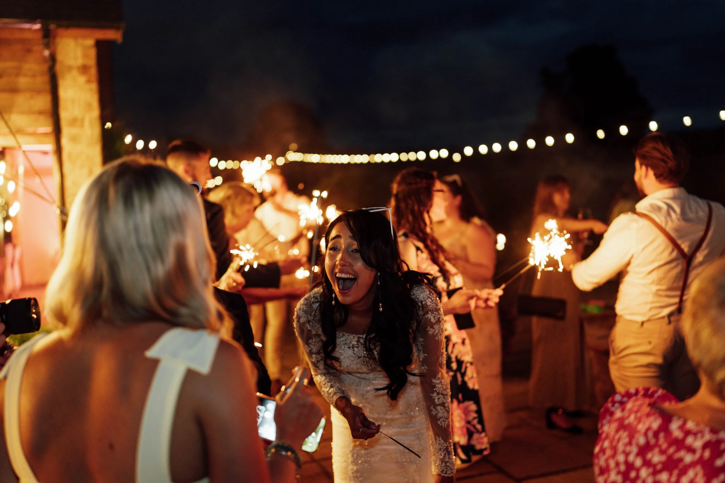 Women and men celebrating at an outdoor nighttime event with sparklers, festive lights, and joyful expressions.