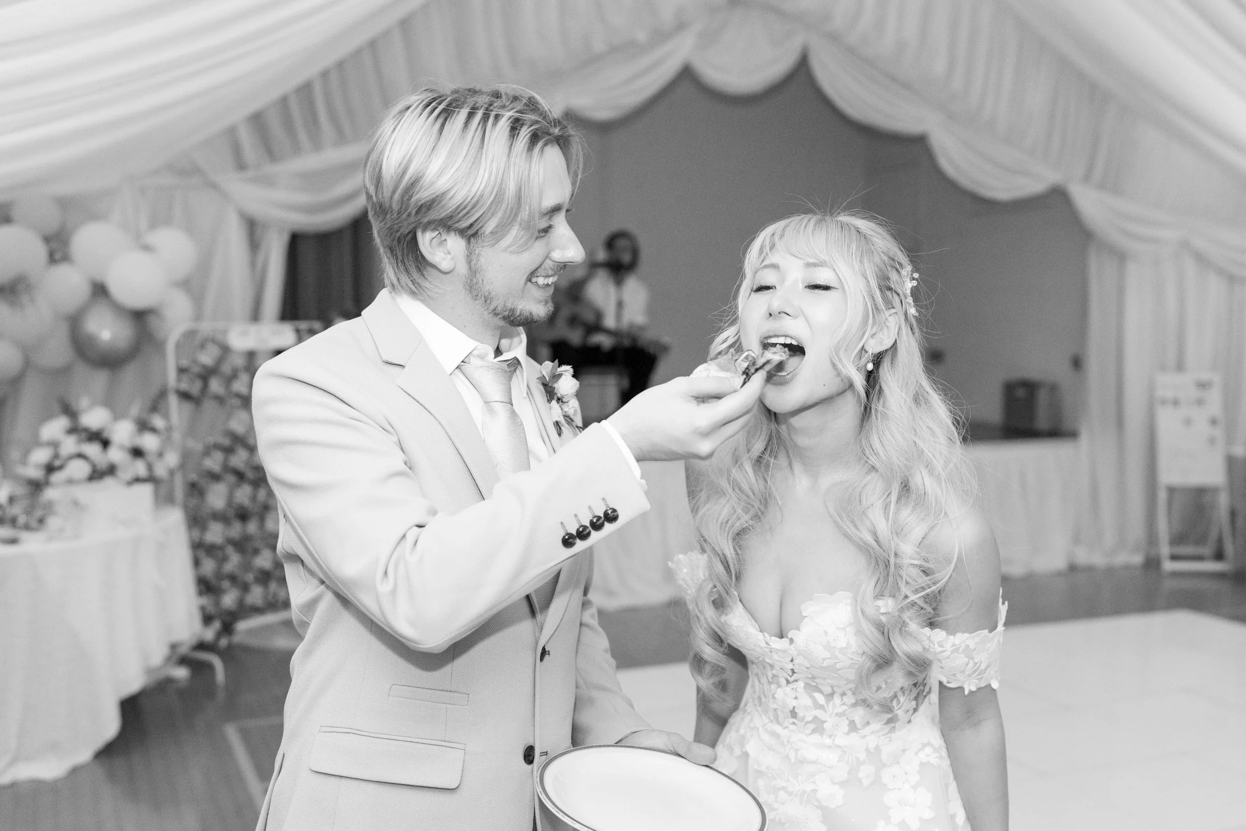 A wedding reception with a groom feeding a bride a piece of cake in a decorated hall.