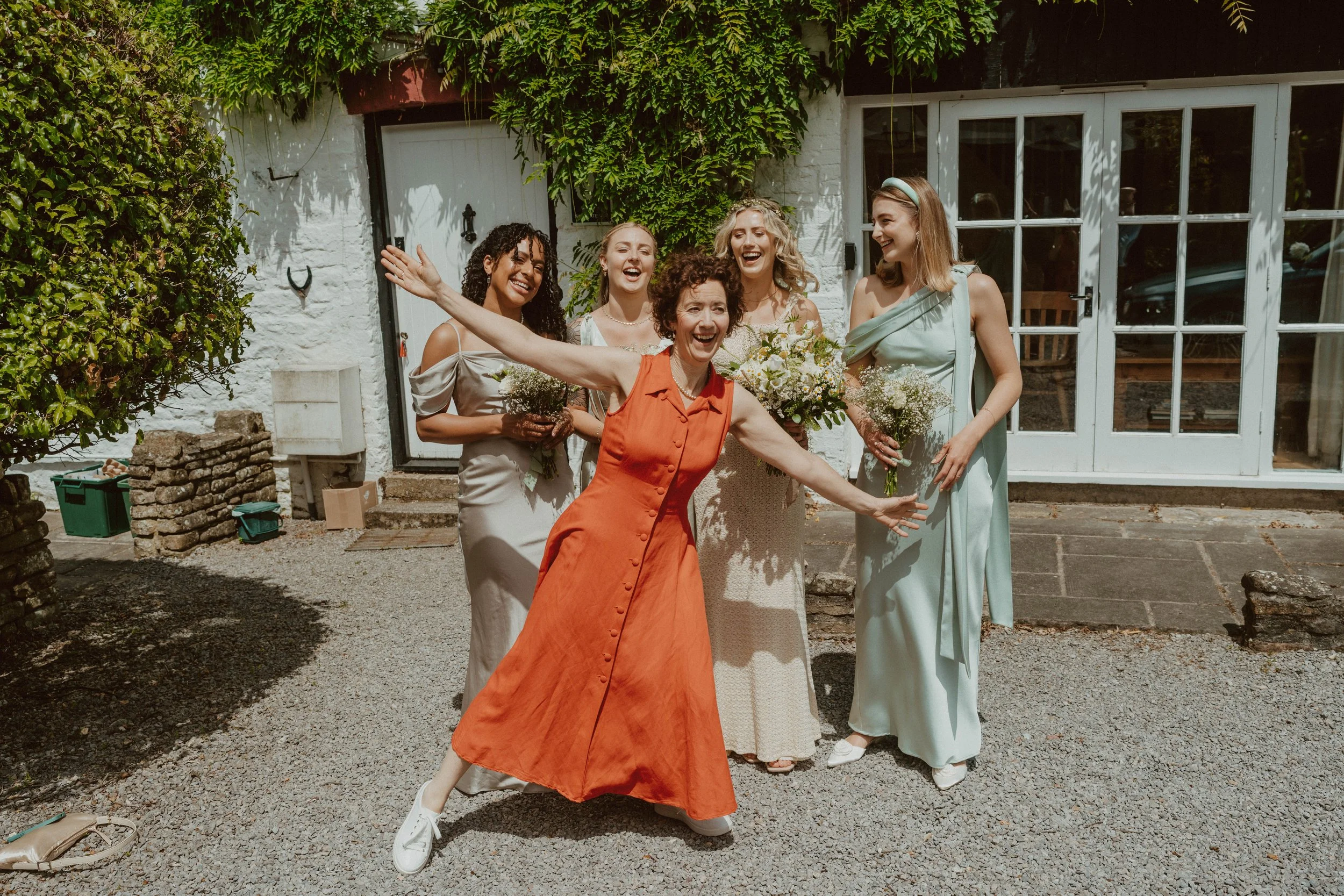 A group of five women celebrating outdoors, holding bouquets of flowers, smiling and laughing on a sunny day.