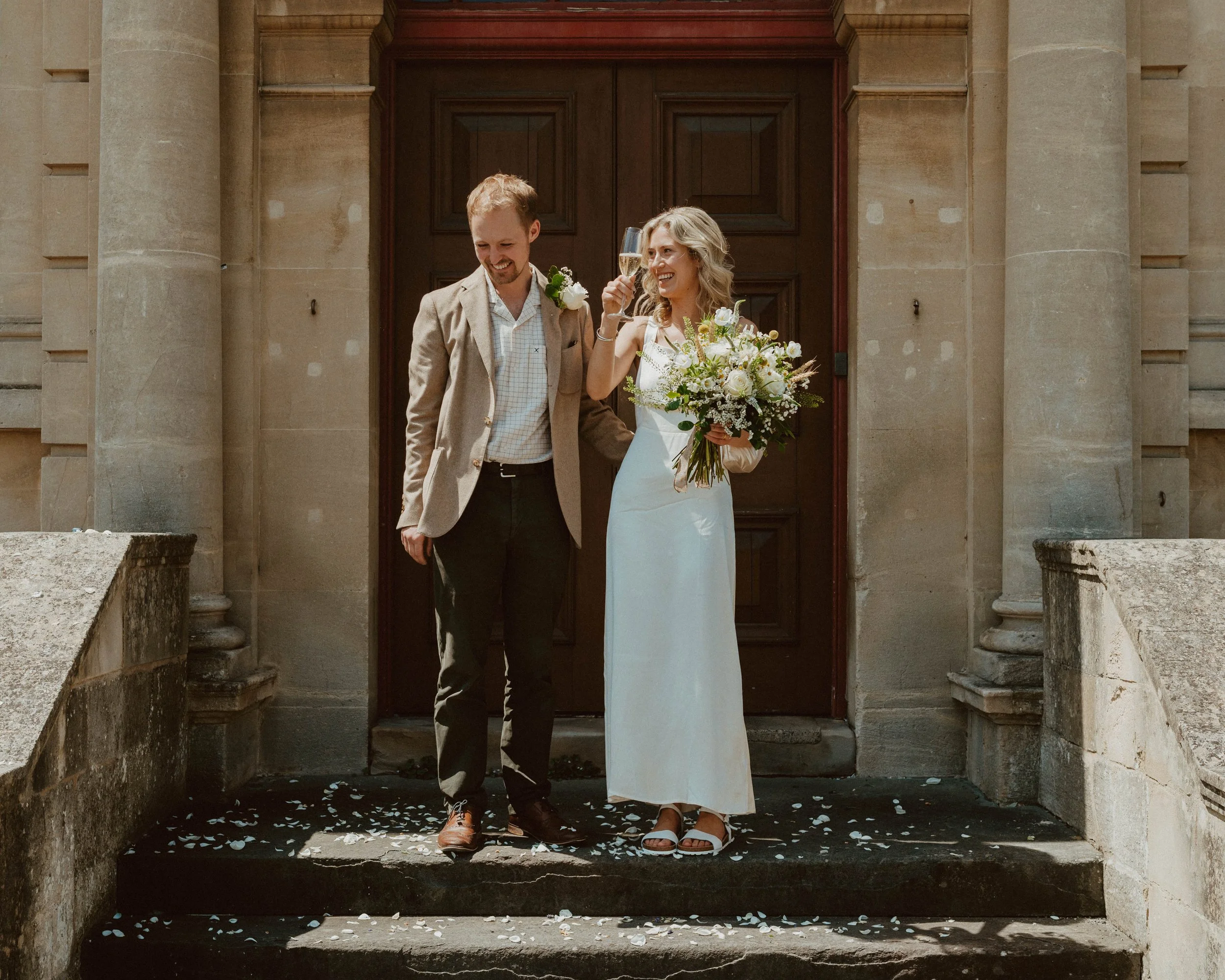 A newlywed couple stands on the steps of a stone building, celebrating with champagne. The bride is holding a floral bouquet, and both are smiling, enjoying their special moment.