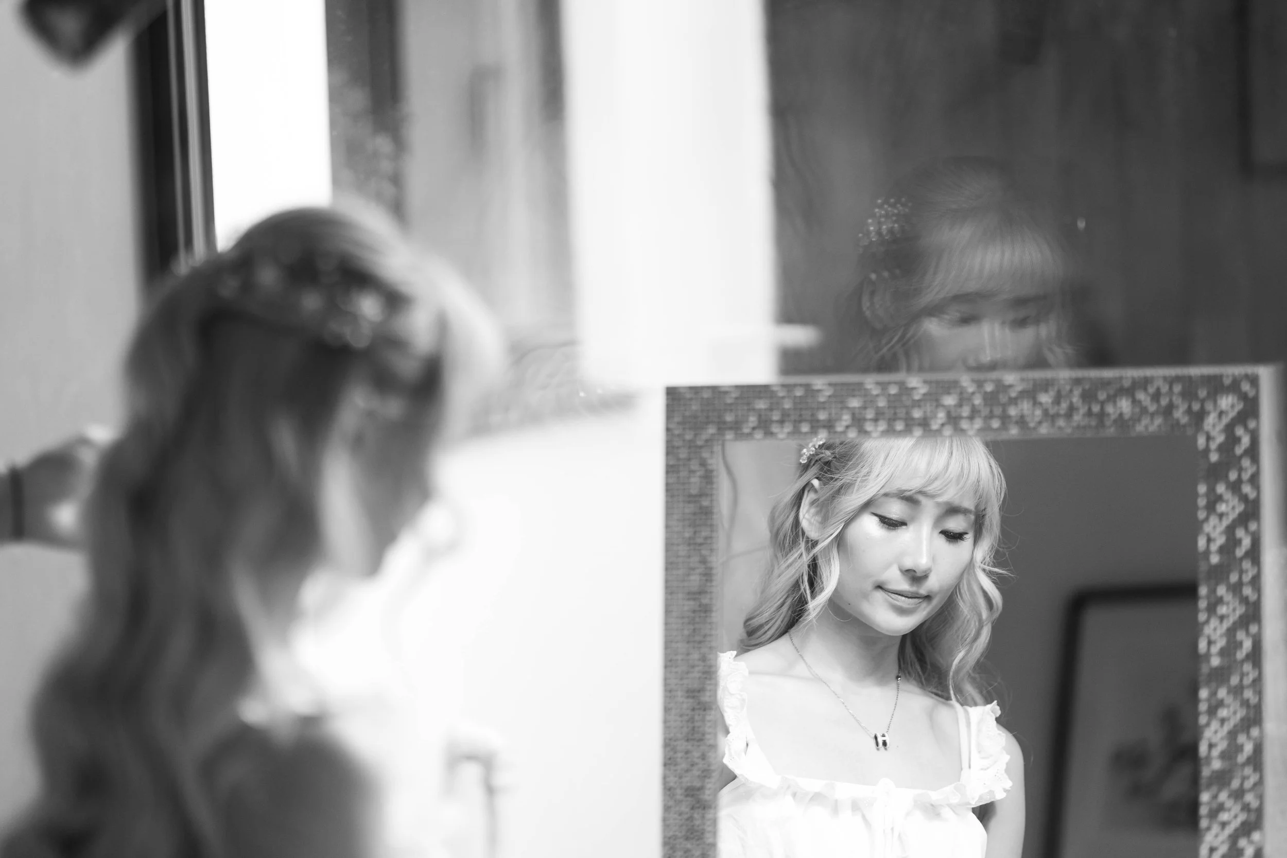 A woman viewed through a mirror, her reflection showing her looking down with a gentle smile, wearing a white dress and a necklace, with her hair styled in loose waves.