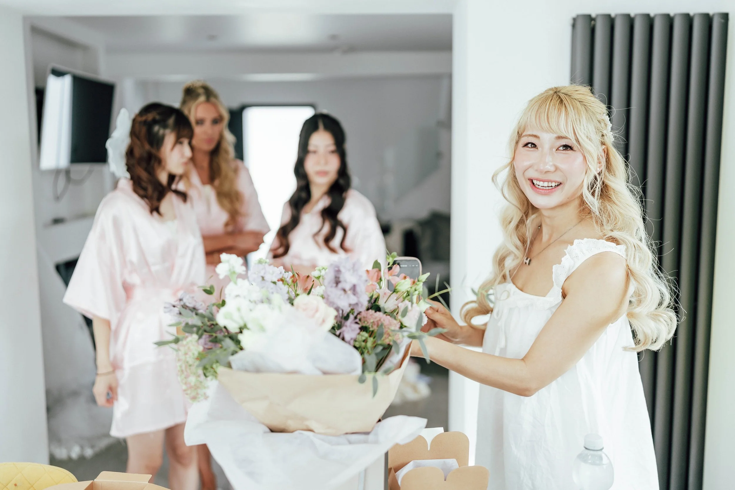 A smiling blonde woman in a white dress takes a photo of a large bouquet of flowers with her phone, while three women in silk robes look on in a bright room.