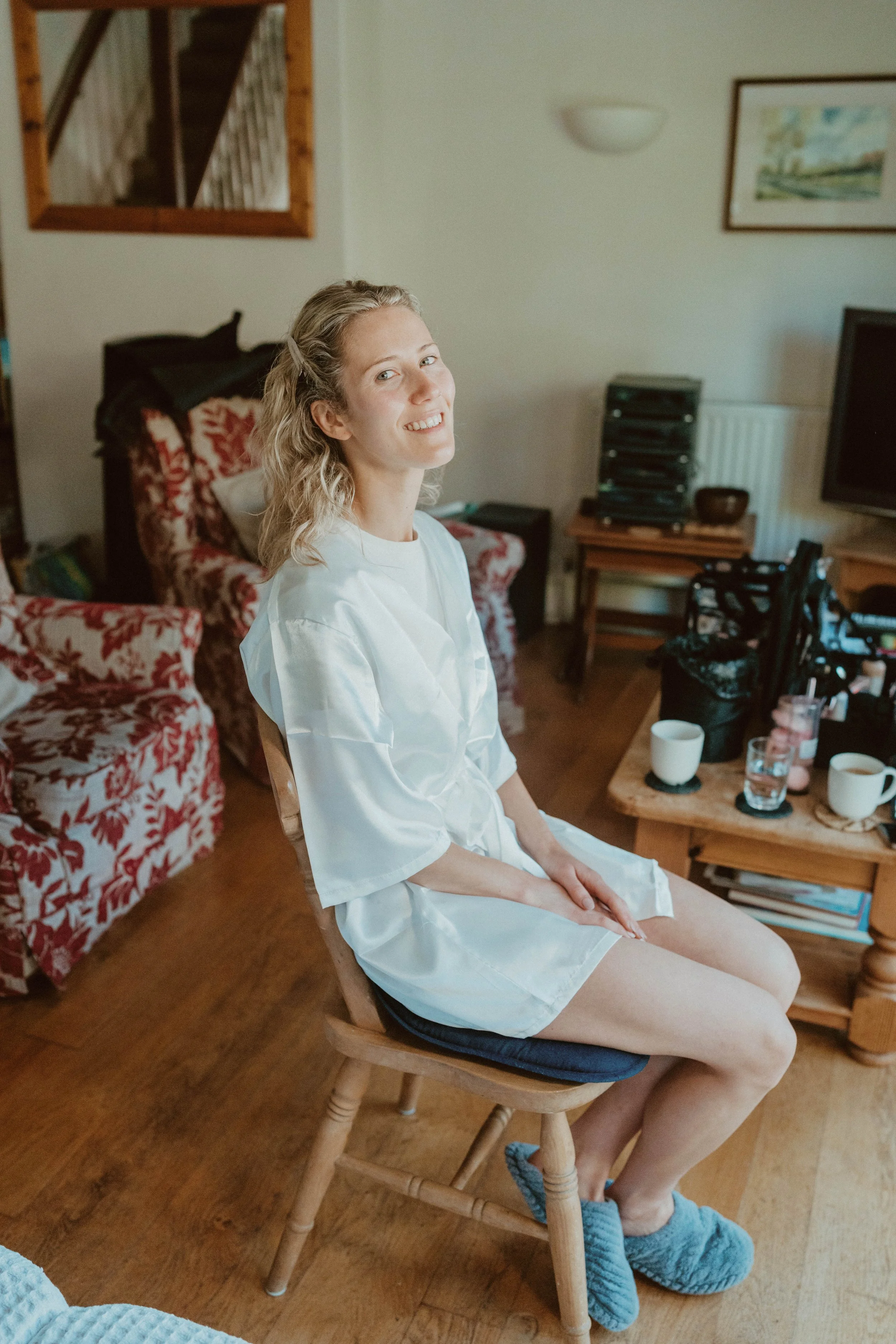 A woman with blonde hair sitting on a wooden chair in a living room, smiling and wearing a white silky robe and gray slippers.