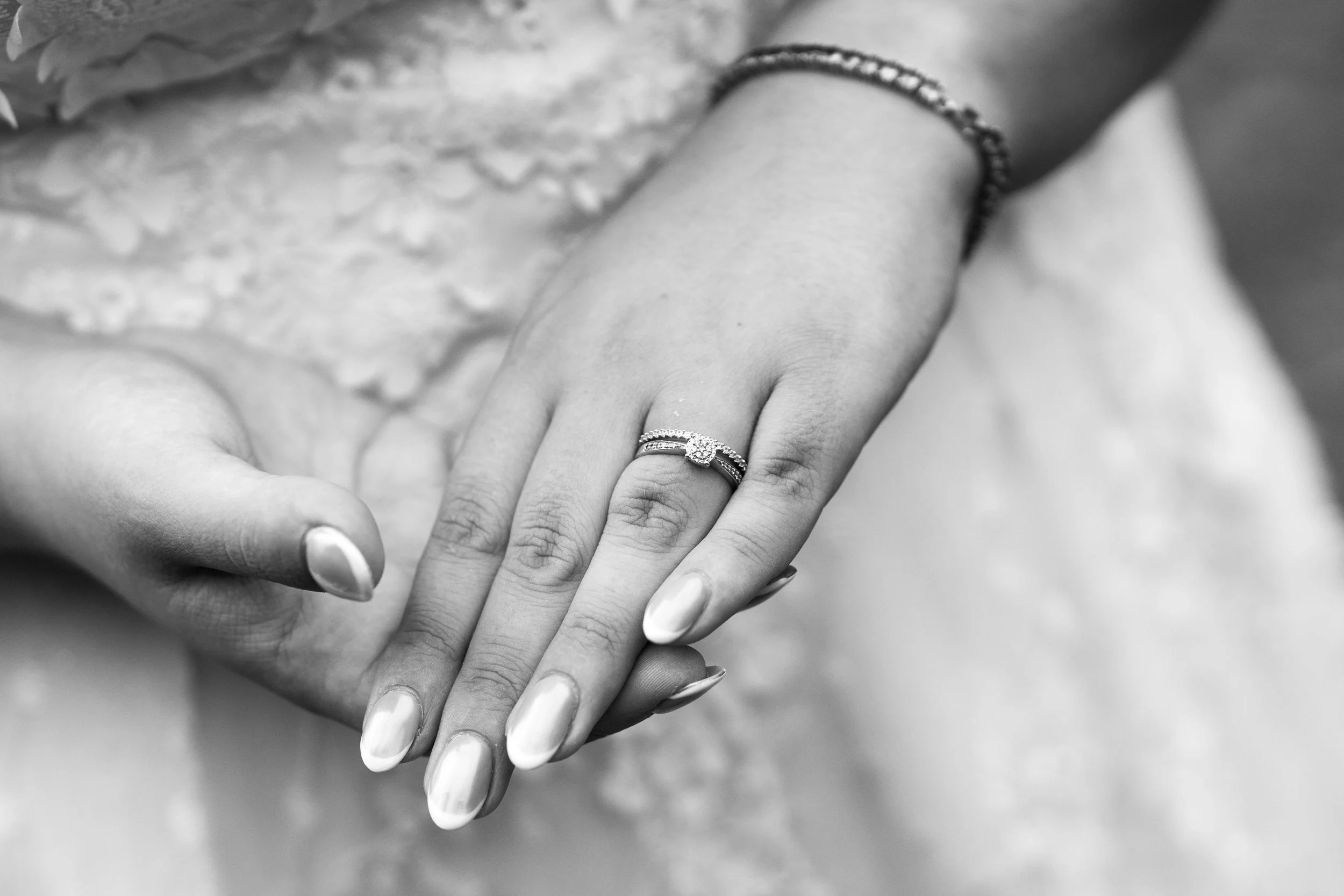 Close-up of two hands, one with an engagement ring with a large center diamond surrounded by smaller stones, and both hands with manicured nails, one wrist with a bracelet.