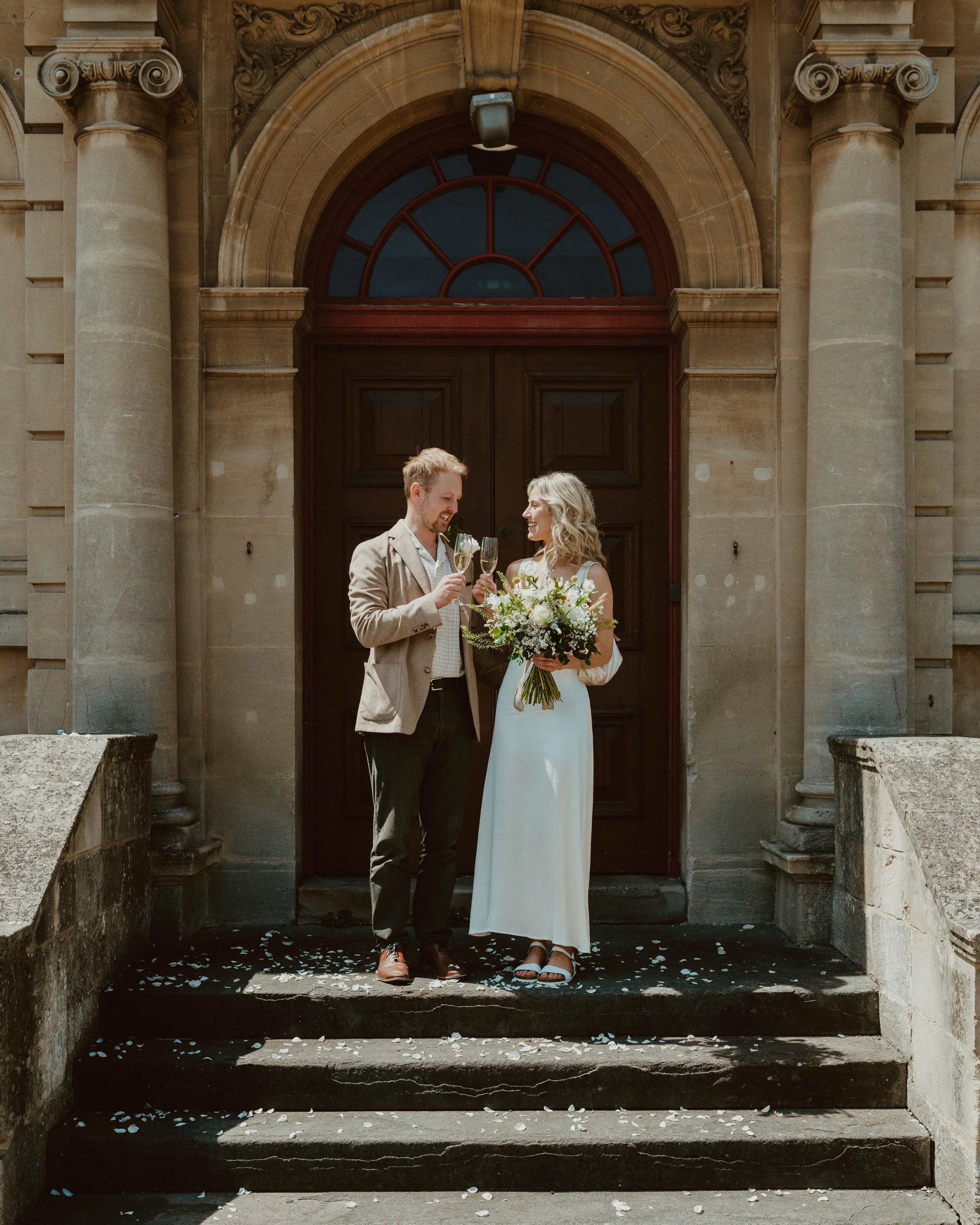 A couple in wedding attire celebrating on steps in front of a stone building with large wooden door, holding champagne glasses and a bouquet of flowers.