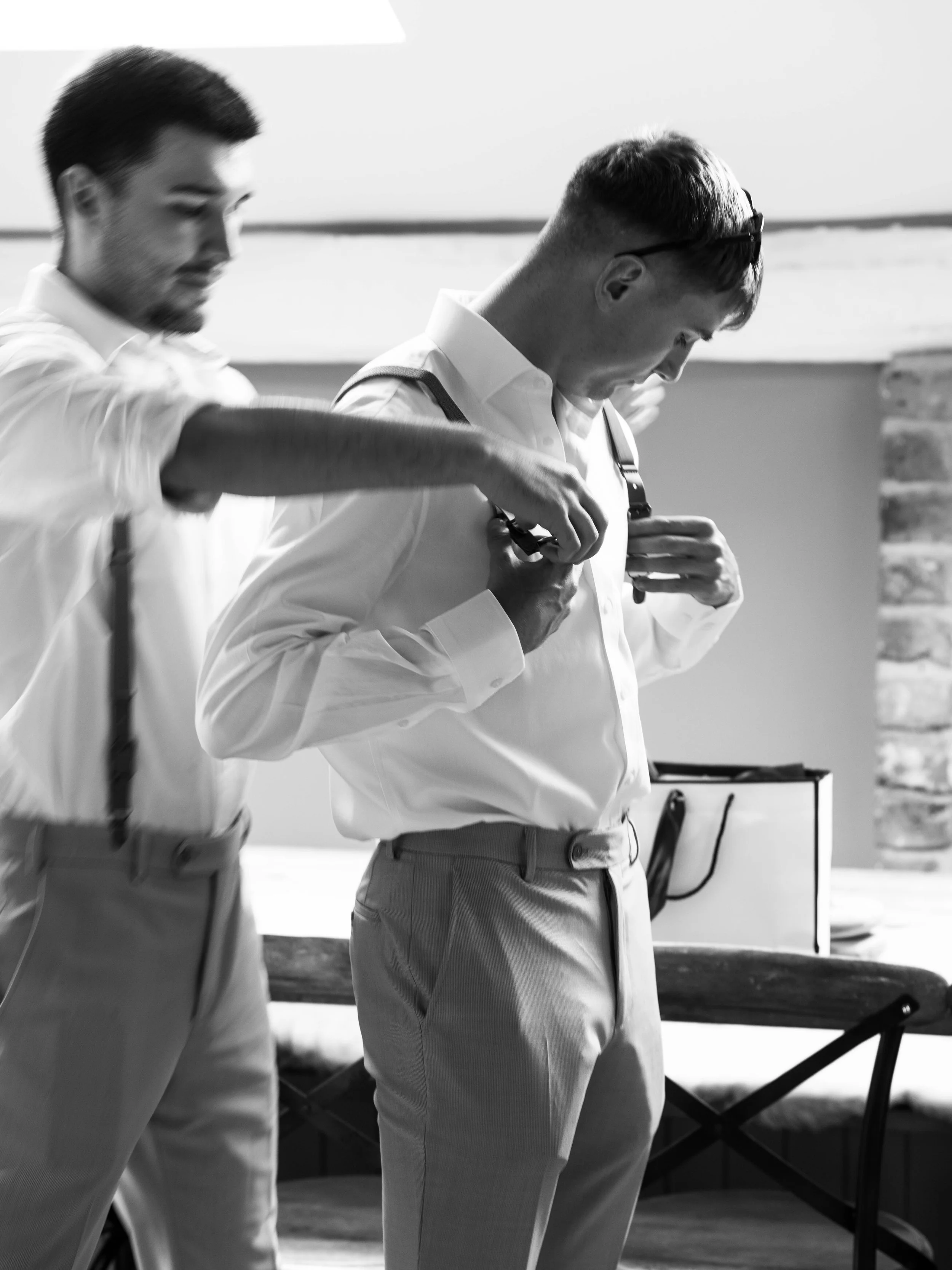 Two men in dress shirts and pants, one assisting the other with suspenders in a room with a table and brick wall.