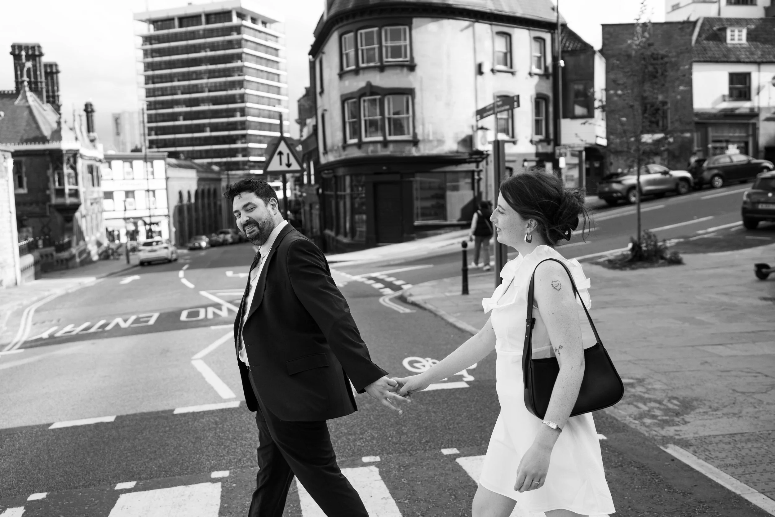 A man in a suit holding hands with a woman in a white dress as they cross a city street, smiling and looking at each other, in an urban neighborhood with tall buildings and cars.