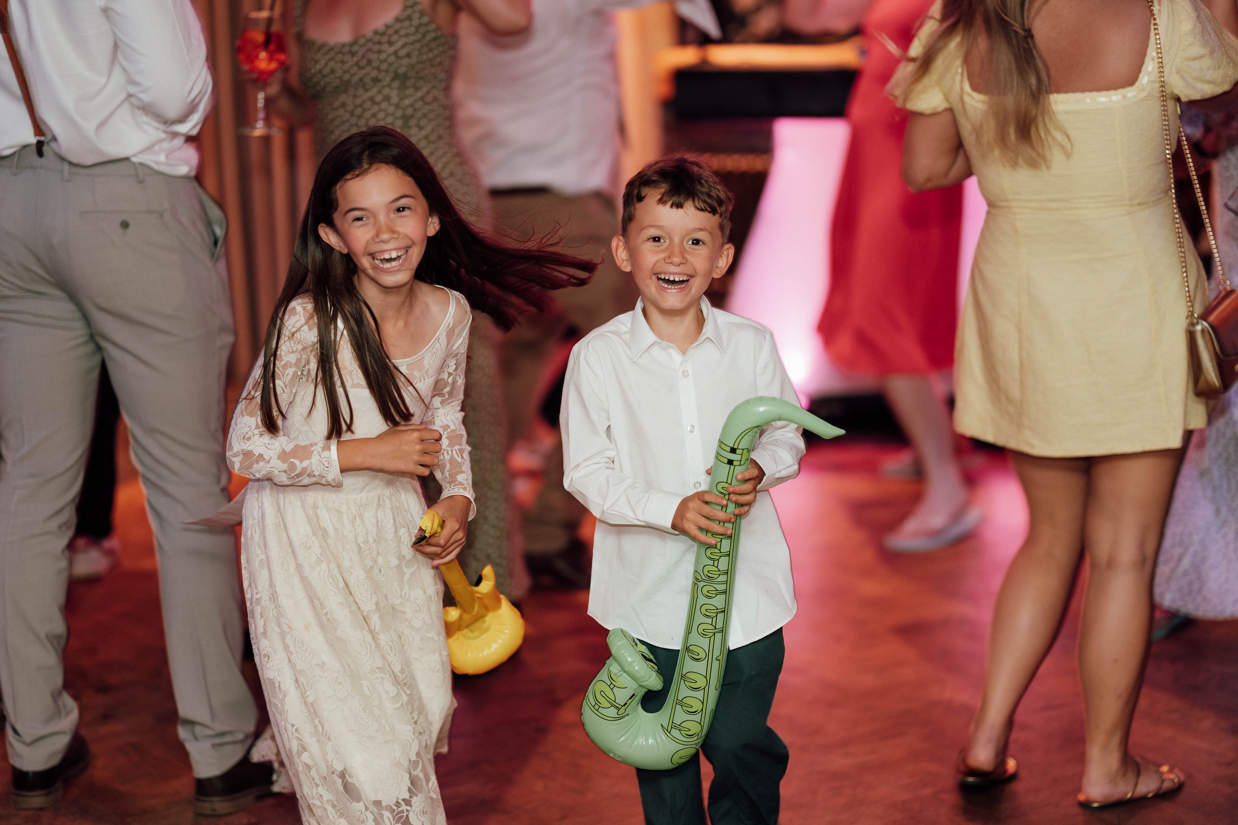 Two children, a girl and a boy, laughing and playing at a party; the boy is holding a green inflatable saxophone, and the girl has a yellow balloon. They are surrounded by adults and other people in casual and semi-formal attire.