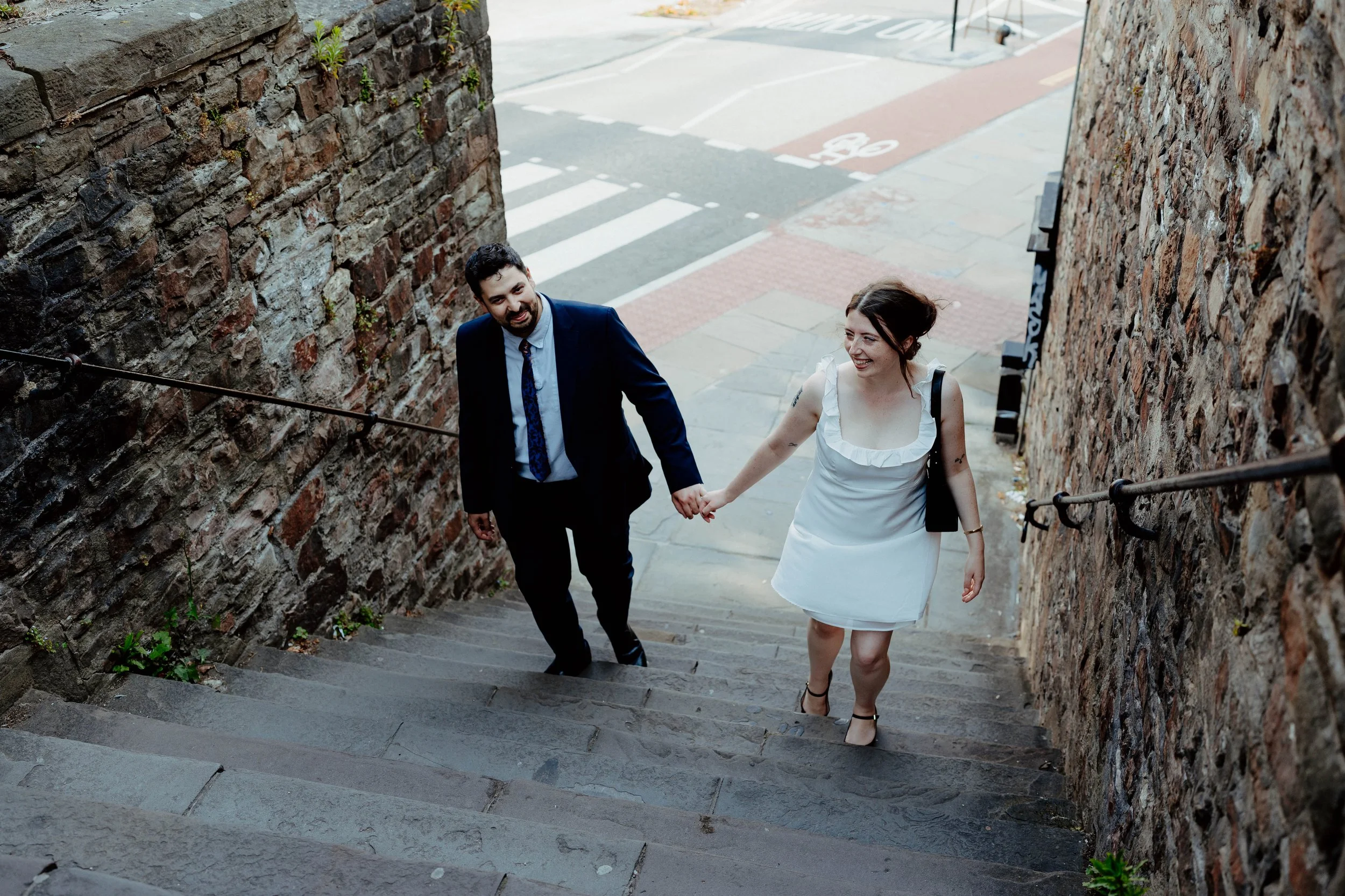A couple holding hands and walking up a stone staircase between brick walls, smiling and looking at each other, with a city street and bike lane in the background.
