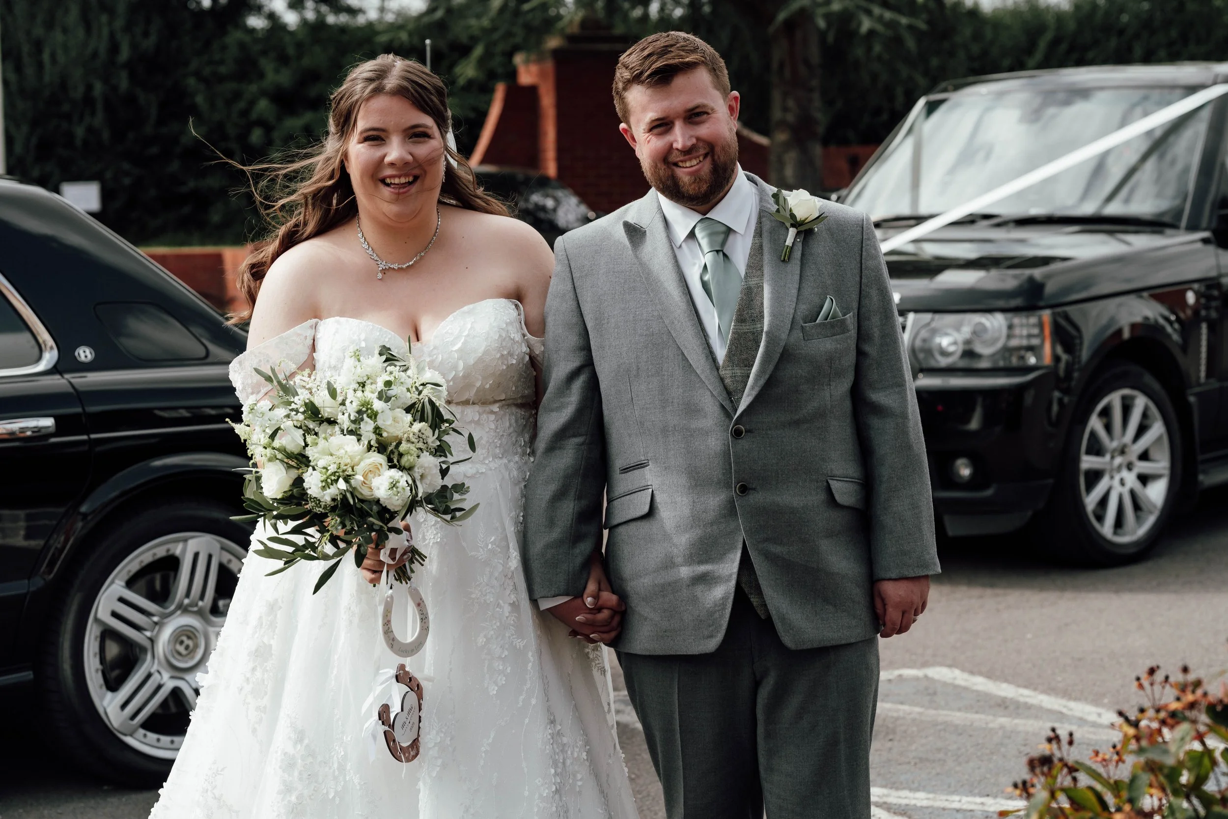 A bride and groom smiling and holding hands outdoors, dressed in wedding attire, with cars and trees in the background.