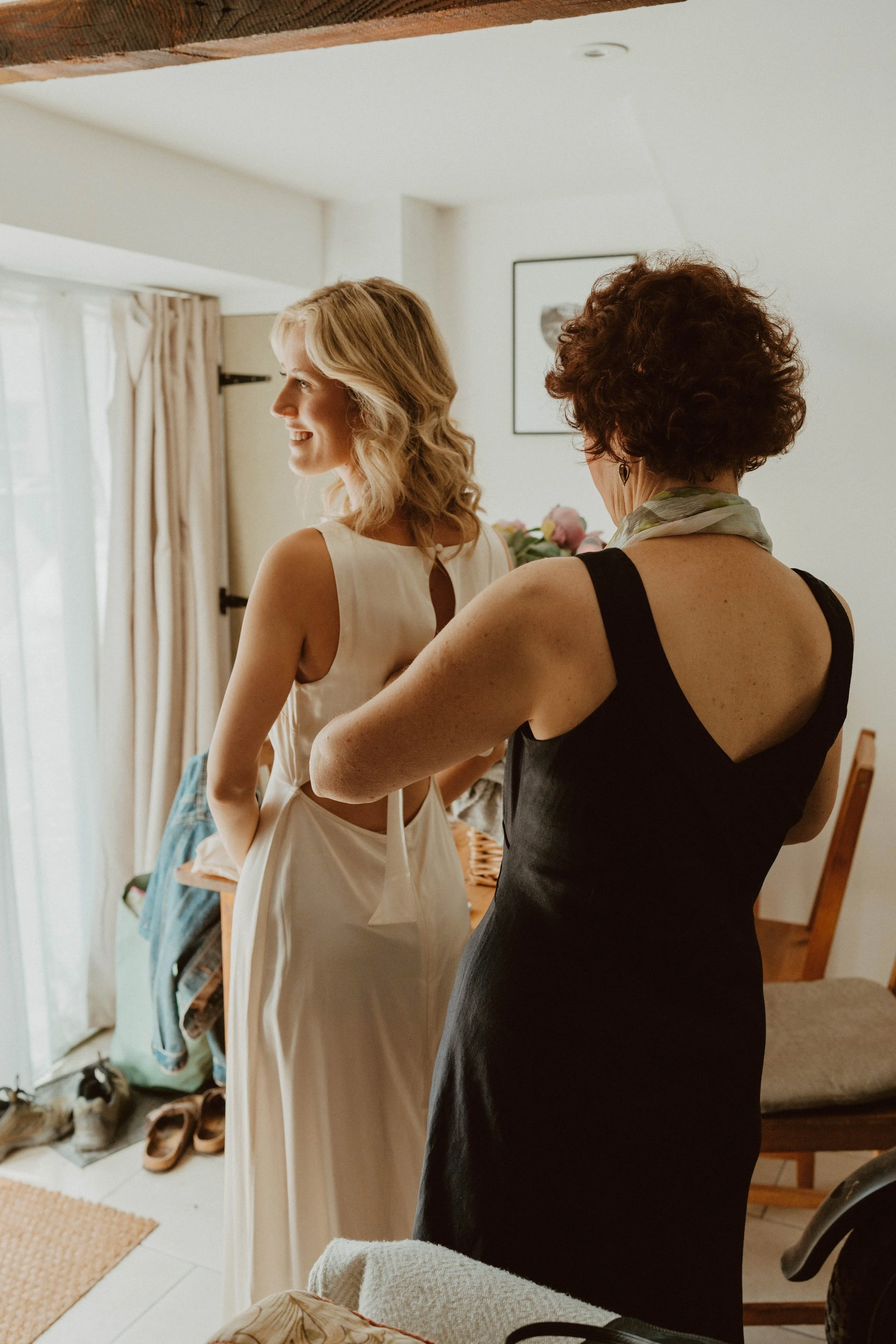 A woman in a white dress getting ready, assisted by another woman in a black dress, inside a cozy room with natural light, curtain, and a small table with flowers.