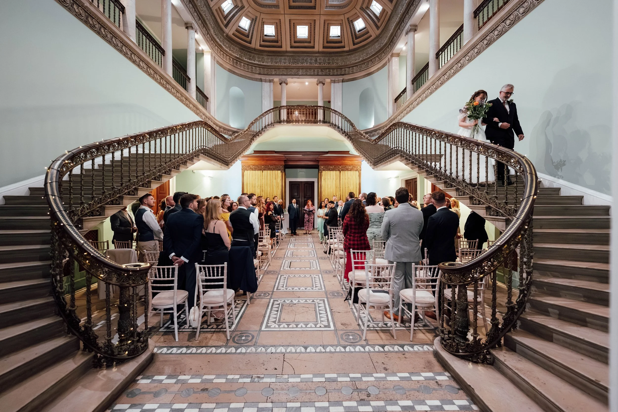 Wedding ceremony in a grand, historic building with a large staircase, where guests are standing in rows facing the front as the bride and groom walk in. The architecture features a high, ornate ceiling, stone columns, and detailed railings.