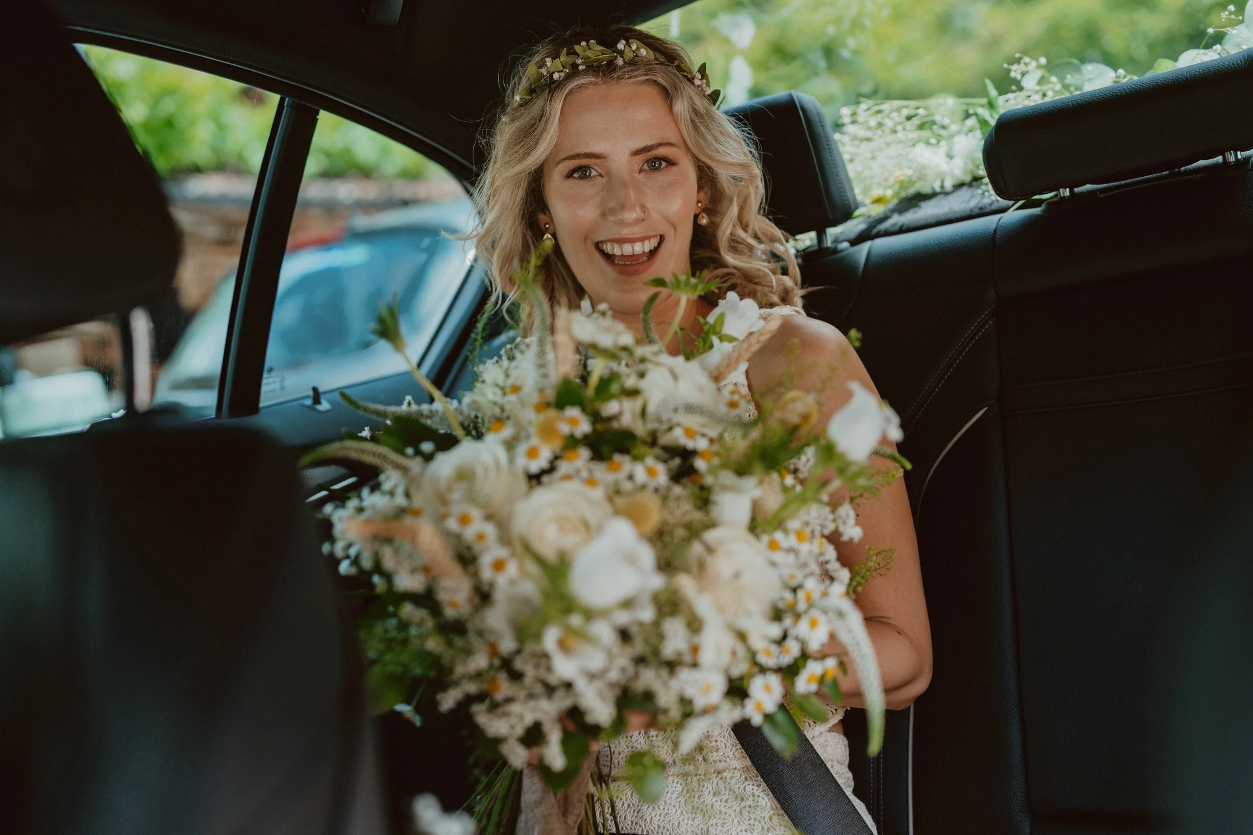 A bride holding a large bouquet of white and yellow flowers inside a car, smiling at the camera with a floral headband and wedding dress.