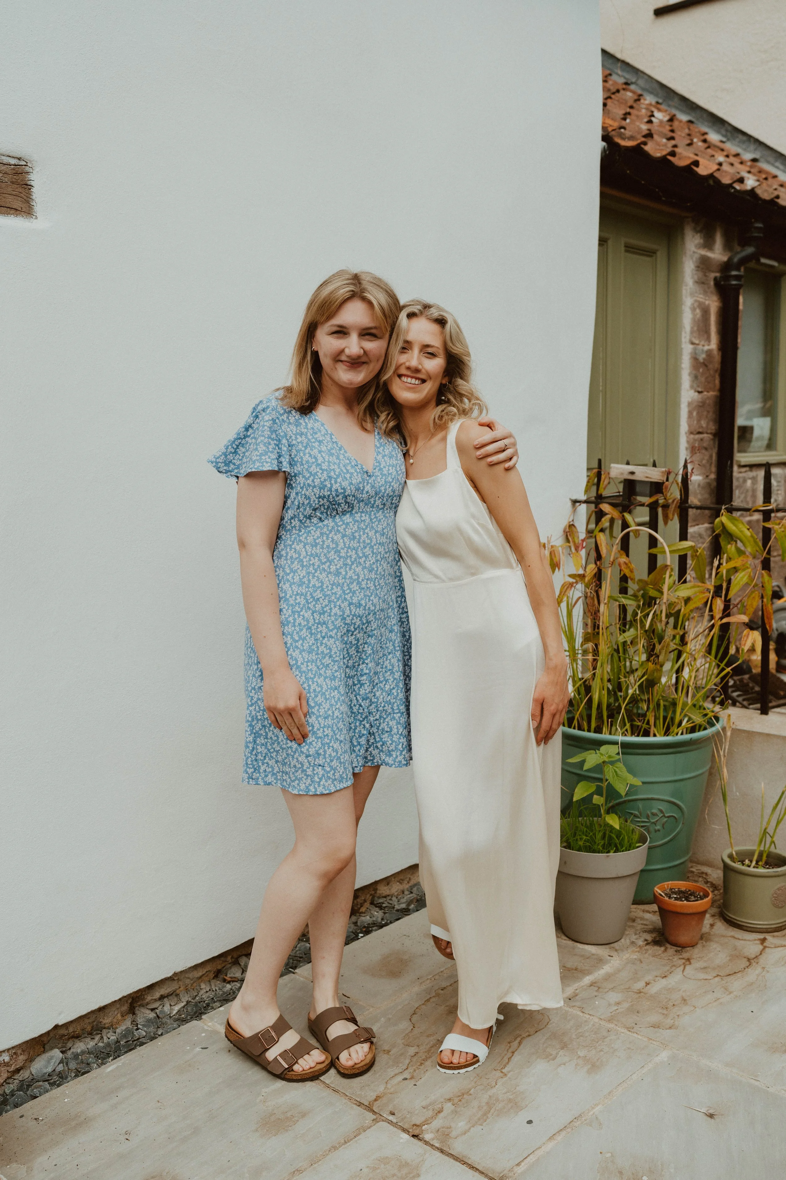 Two women smiling and hugging in an outdoor setting with potted plants and a white wall.