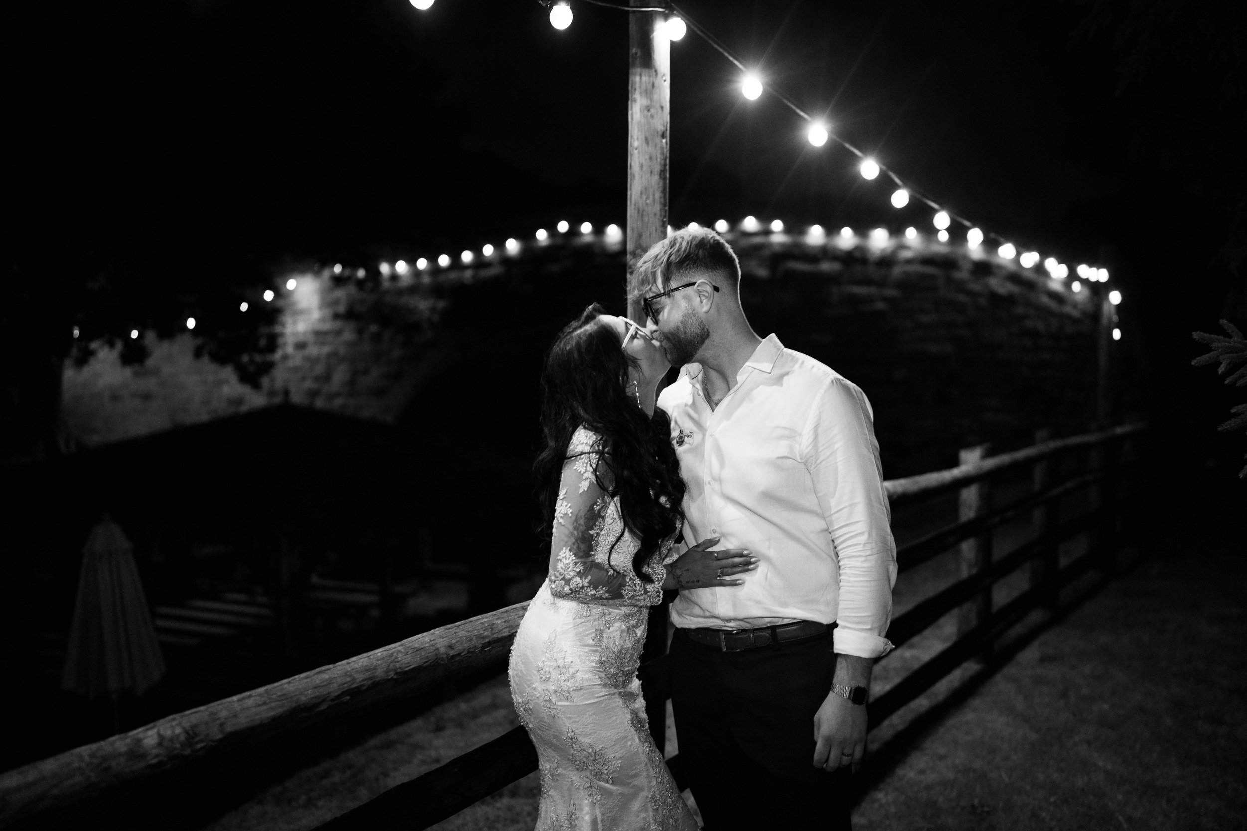 A couple sharing a kiss at night outdoors, illuminated by string lights overhead, with a wooden fence in the background.
