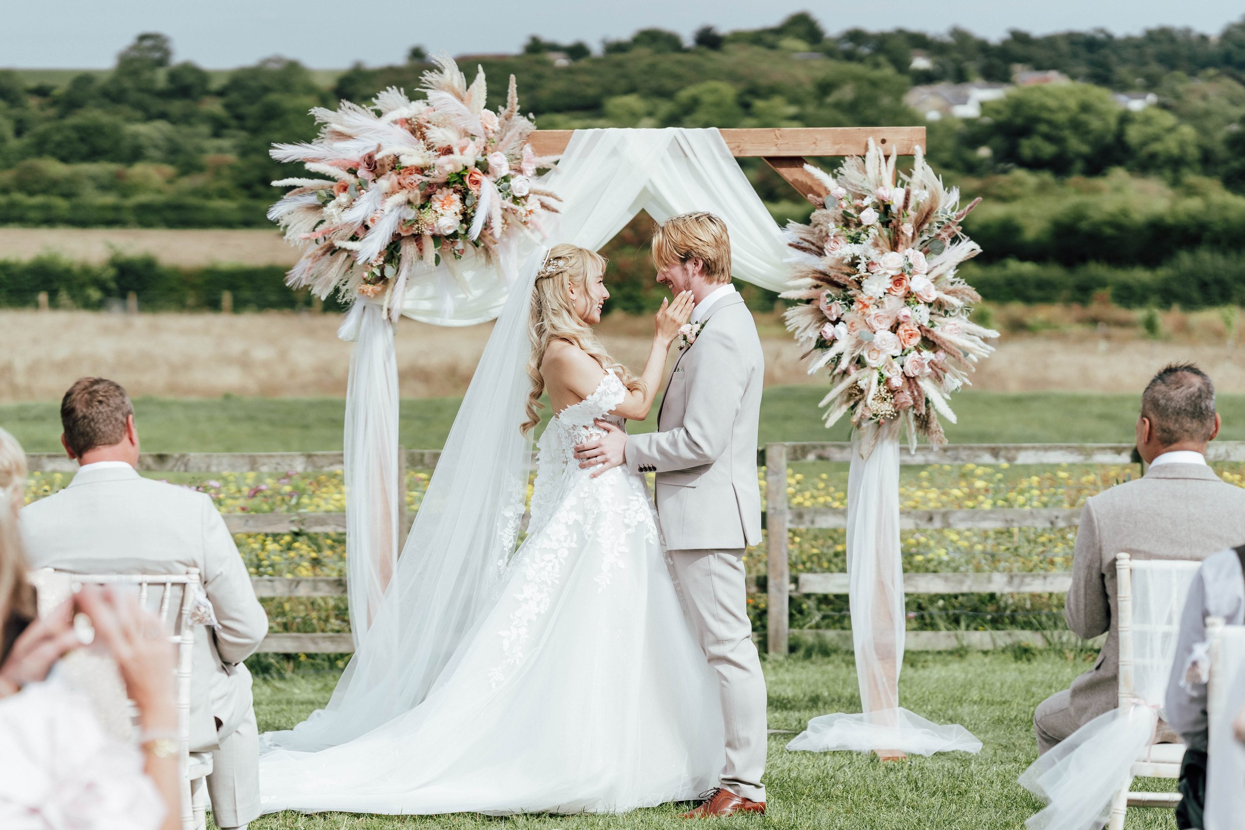 A bride and groom sharing a kiss at an outdoor wedding ceremony, with guests seated nearby and a floral wedding arch behind them.