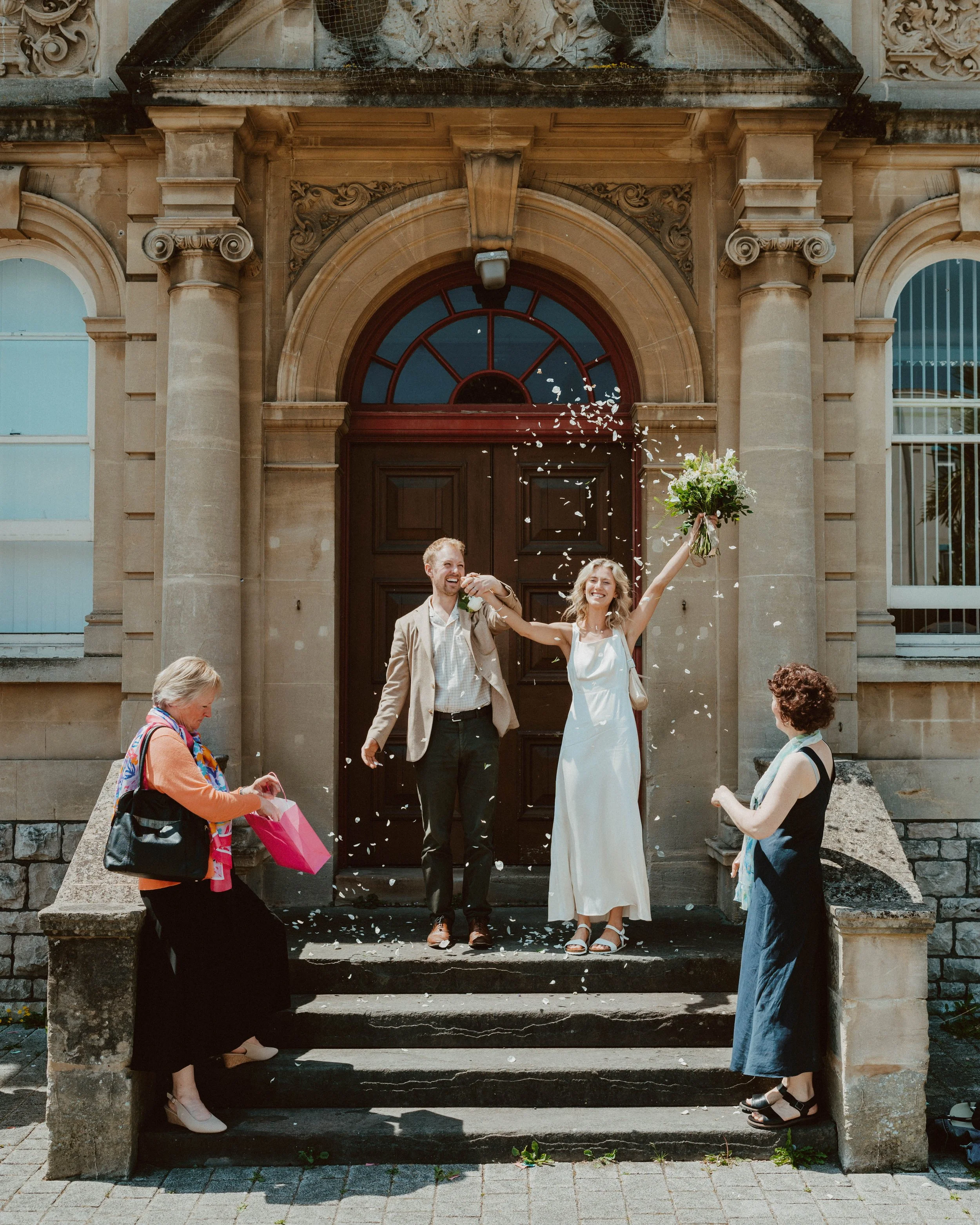 A newlywed couple celebrating on the steps of a building, with flower petals being thrown in the air. The bride is holding a bouquet and smiling, while the groom stands beside her. Two women, possibly family or friends, are nearby, with one holding a