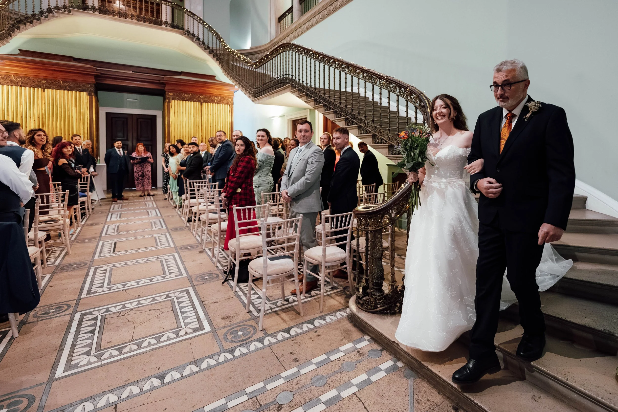 Bride walking down the aisle with her father in a grand, historic wedding venue, with friends and family standing and seated nearby.