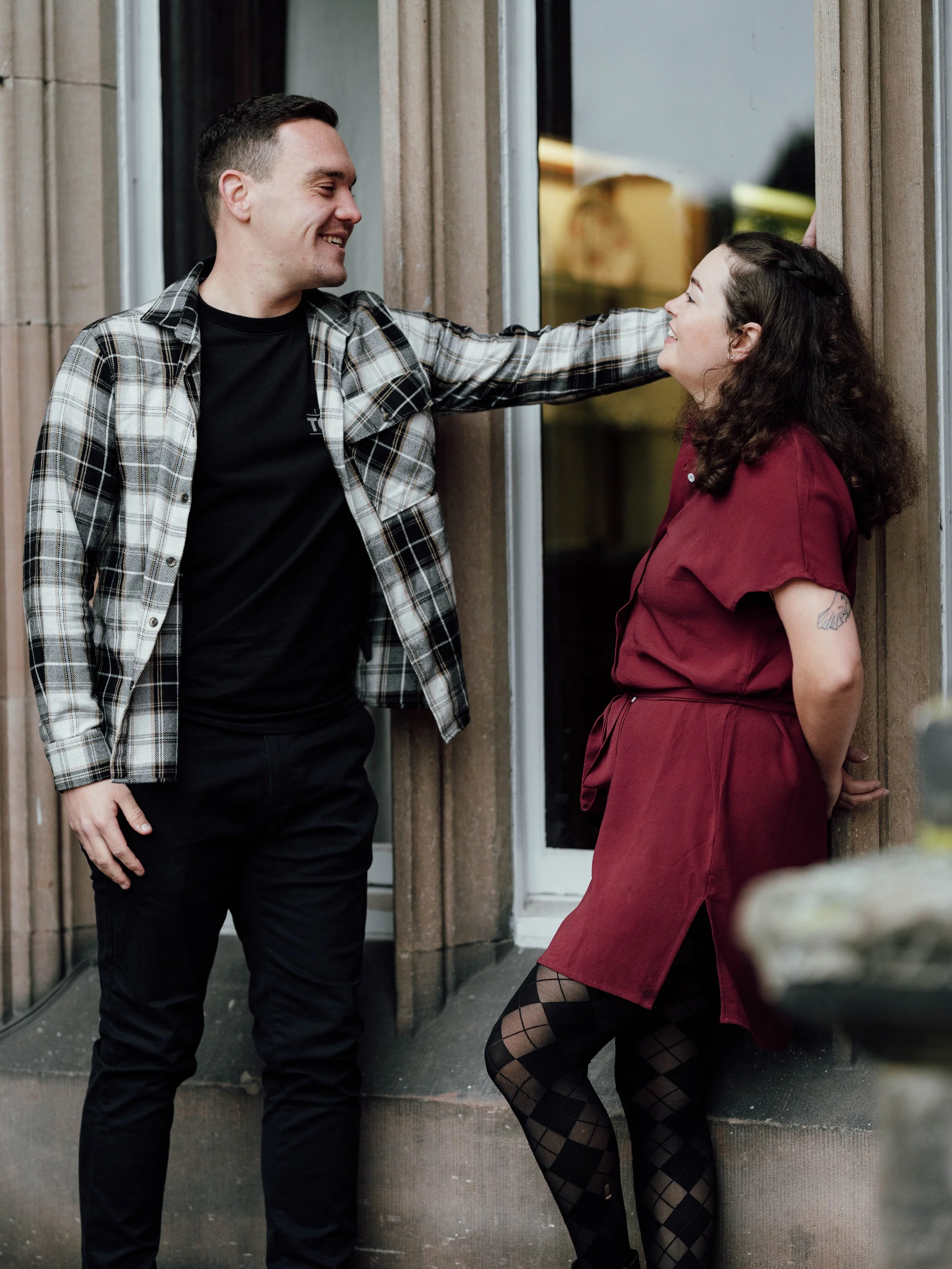 A man and woman living inside a building, standing on the steps outside a glass door, smiling at each other, with the man leaning on the doorframe.