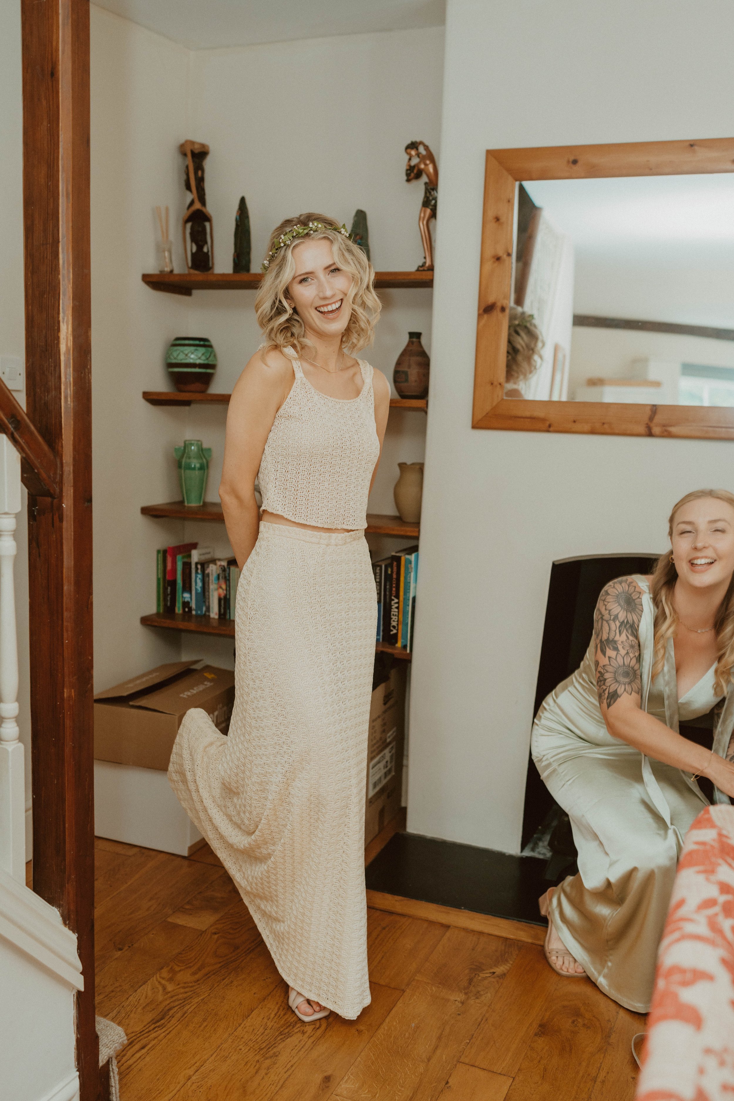 A woman smiling and standing with her hands behind her back, wearing a cream-colored crop top and long skirt, with another woman sitting beside her near a mirror, both in a cozy room decorated with books, vases, and wooden accents.