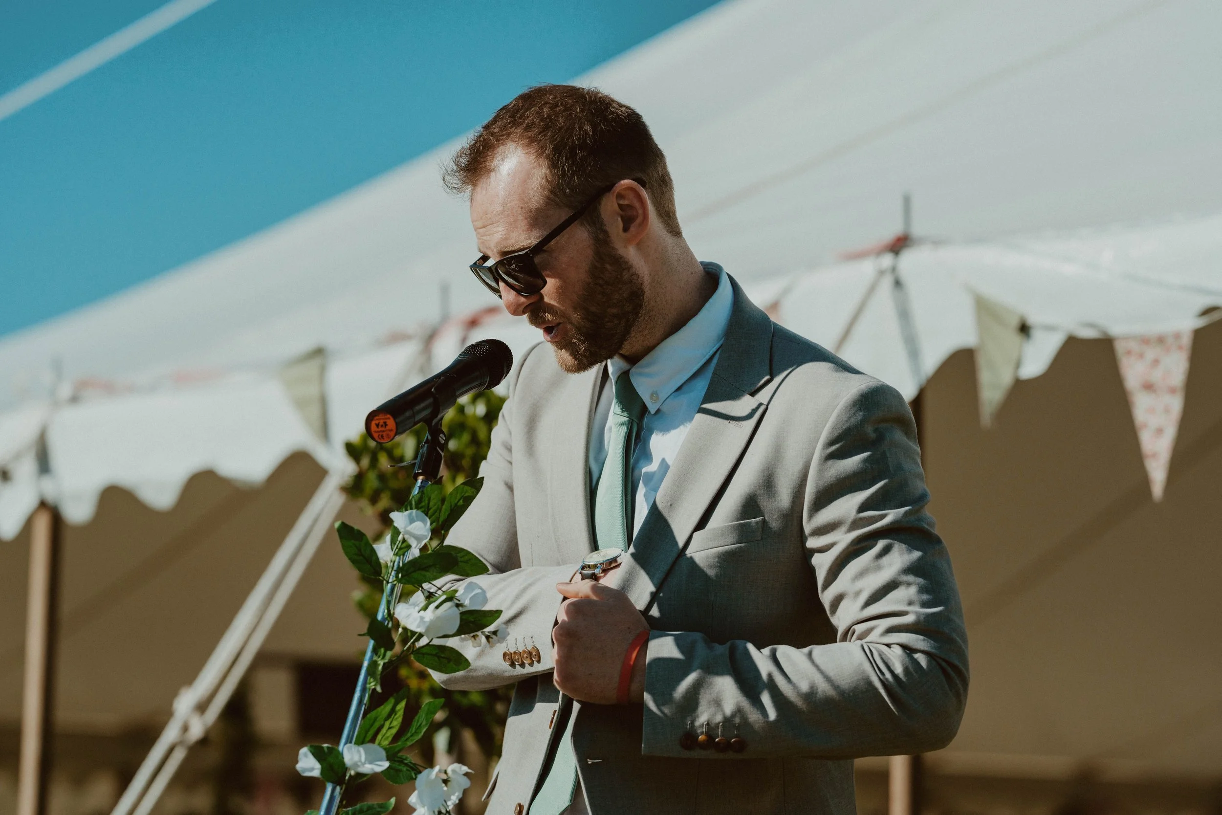 A man in a light gray suit and sunglasses is speaking into a microphone, holding a watch, with white umbrellas and bunting in the background.