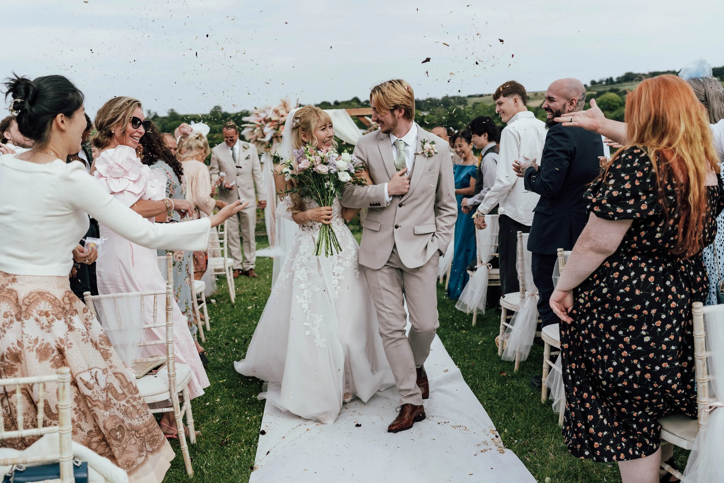 Bride and groom walking down the aisle at an outdoor wedding, surrounded by guests applauding and celebrating.