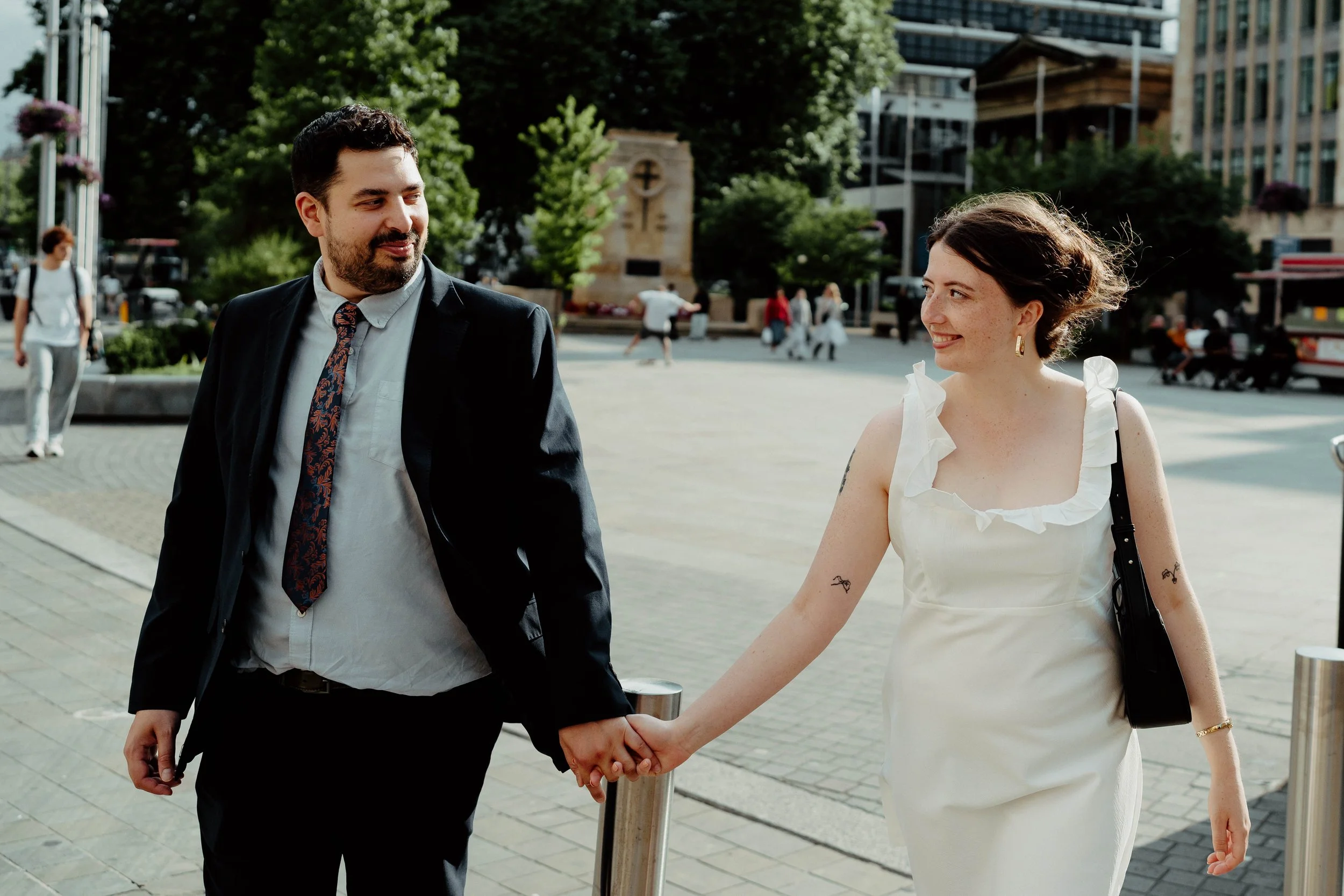A man and woman holding hands and walking in an urban area with trees and buildings in the background, smiling at each other.