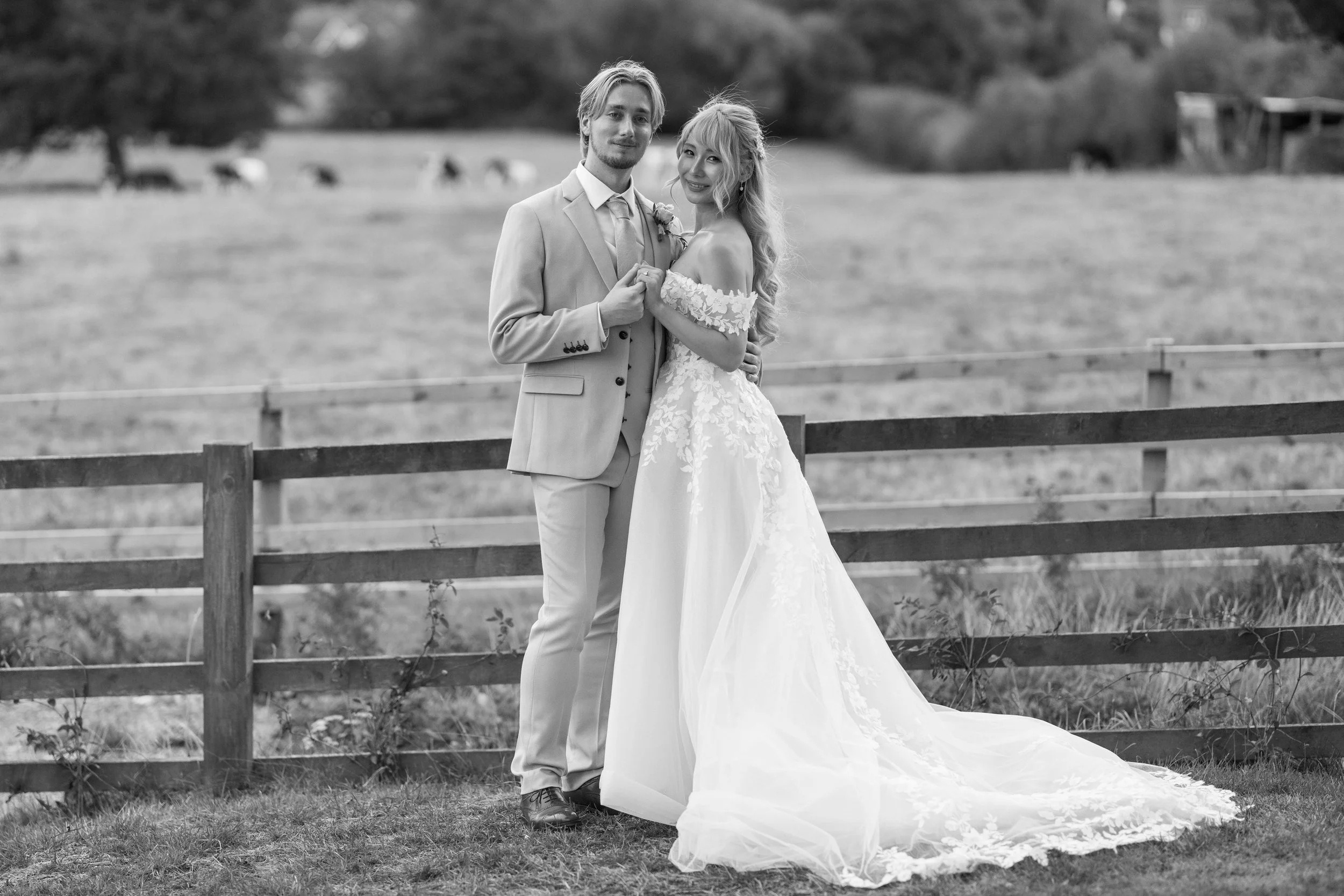 A bride and groom stand together outdoors behind a wooden fence, dressed in wedding attire, with a field and trees in the background.