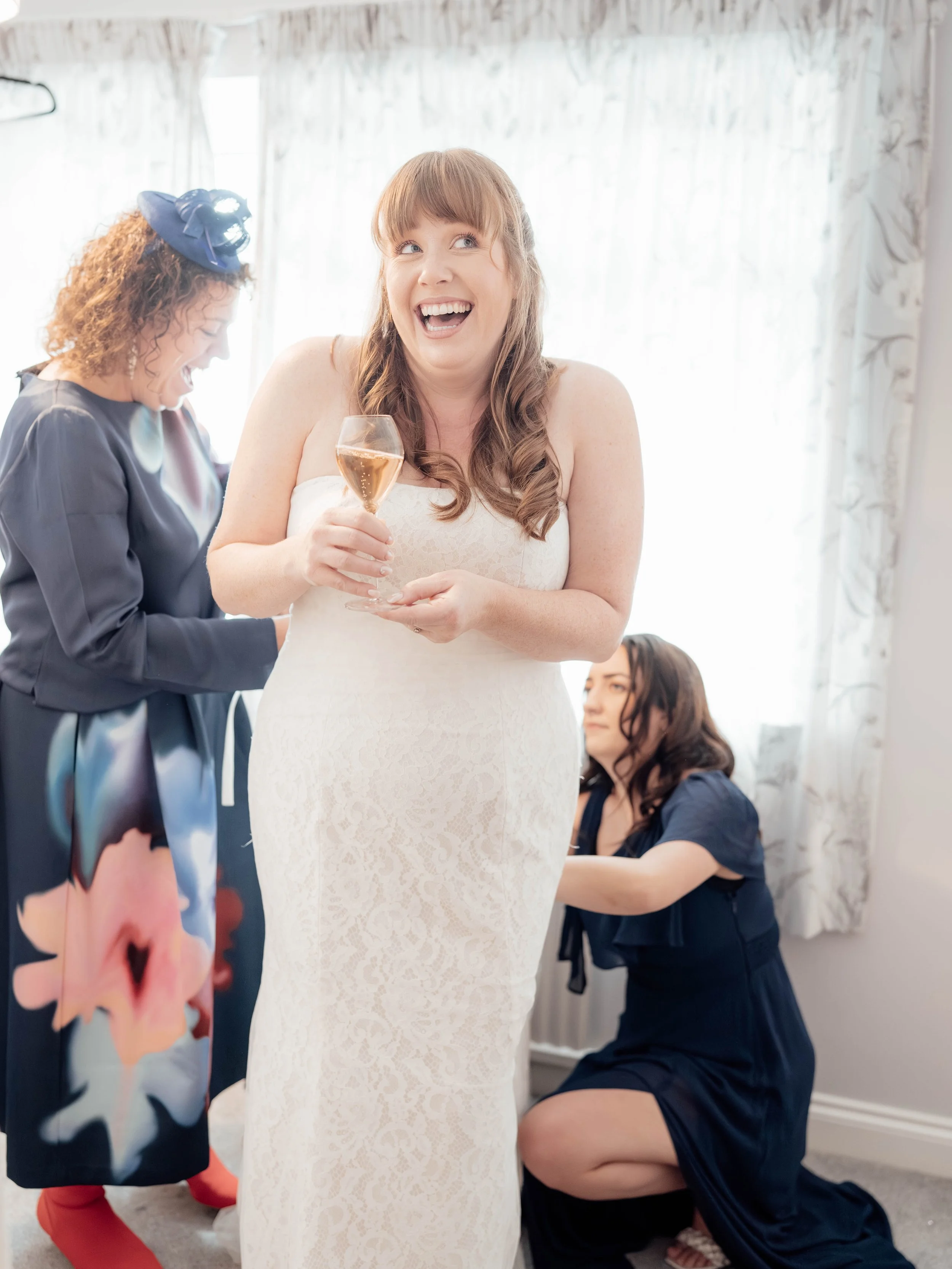 A woman in a white lace dress holding a glass of champagne, smiling and laughing, with two women in navy dresses helping her get ready.