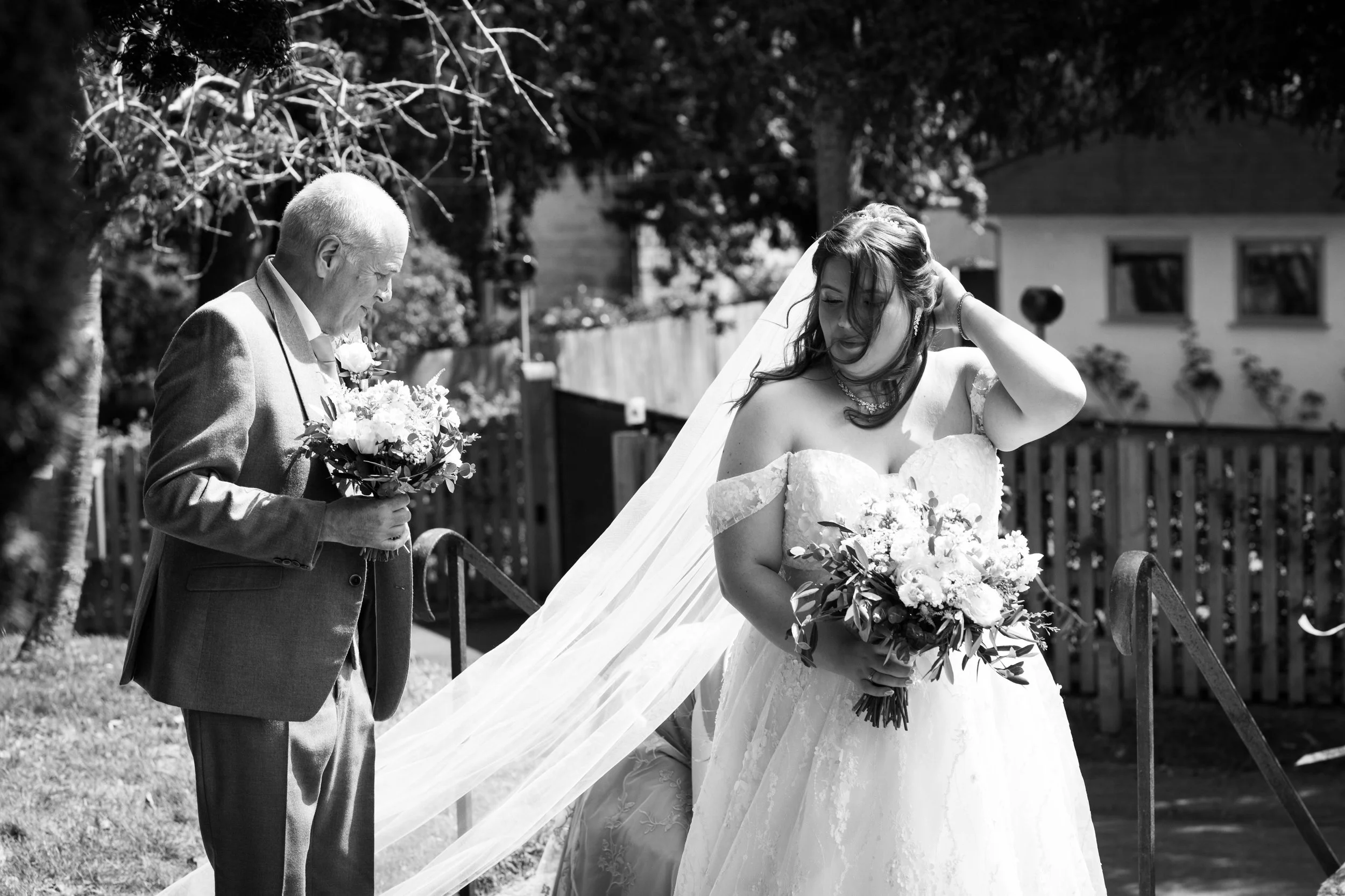 A bride in a wedding dress holding a bouquet of flowers, with an older man in a suit holding a similar bouquet, outdoors on a sunny day.
