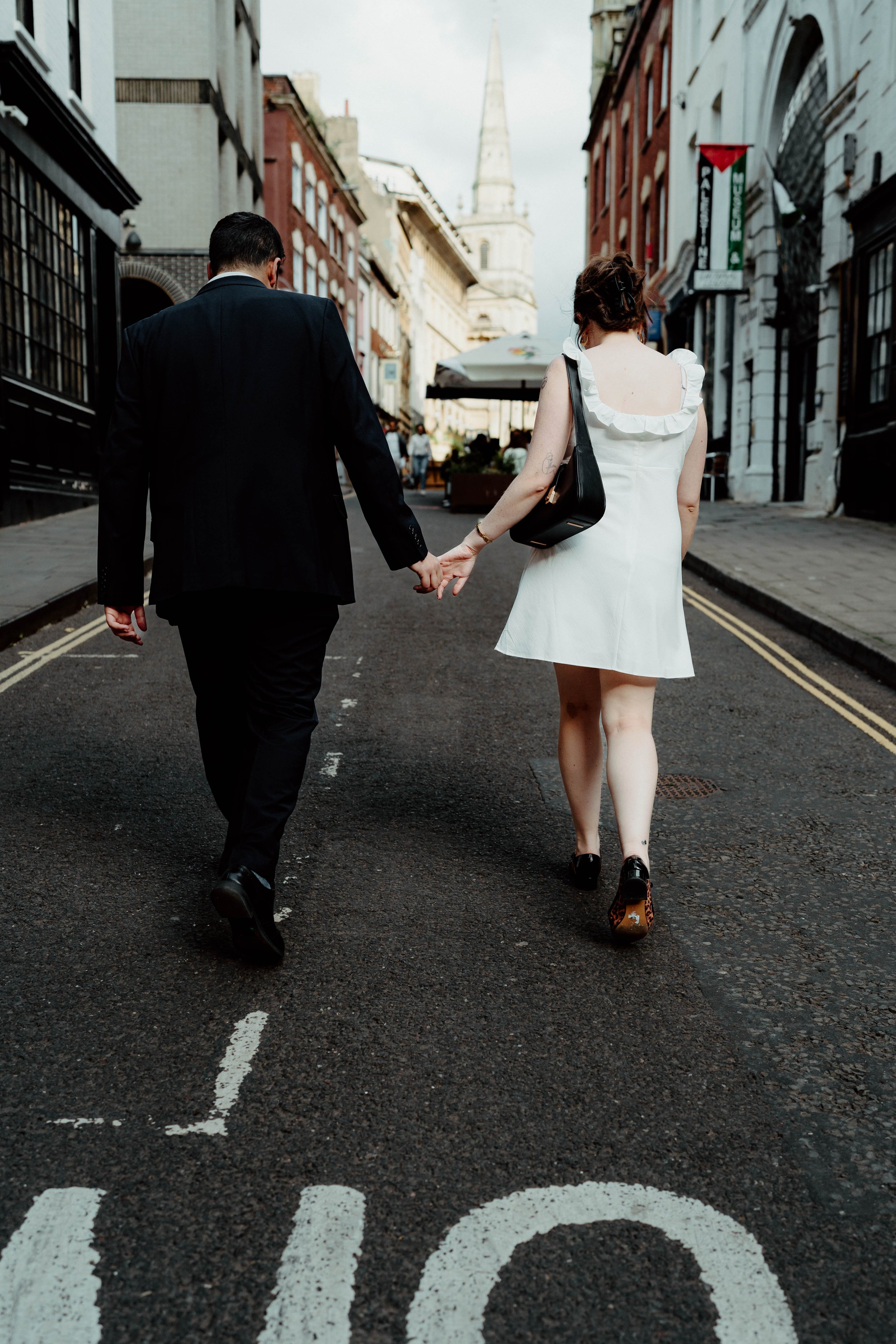 A couple walking hand in hand down a city street, with the woman in a white dress and man in a black suit. The street has buildings on both sides and a church steeple in the background.