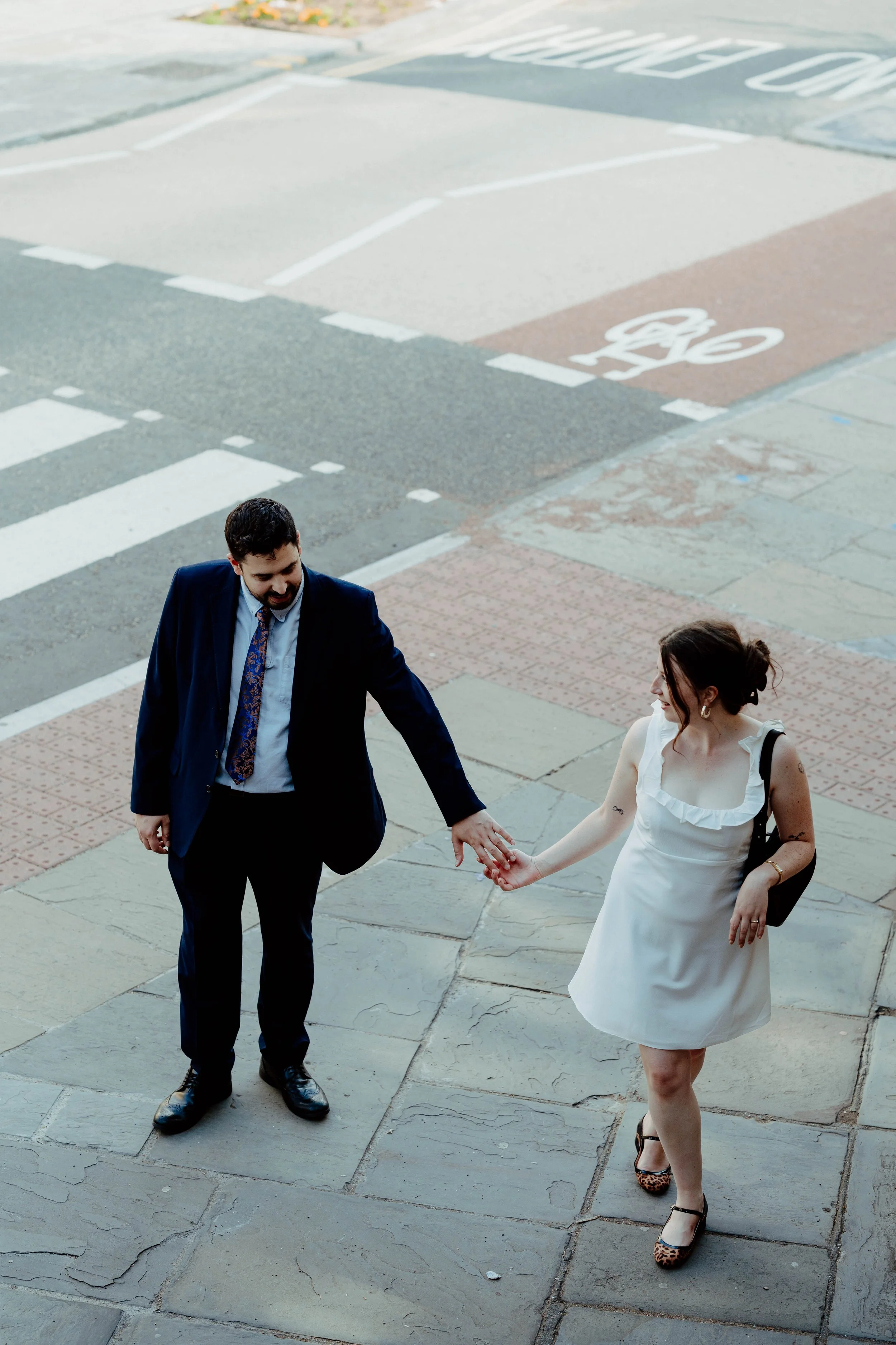 A man in a suit and a woman in a white dress holding hands on a sidewalk near a crosswalk and bike lane