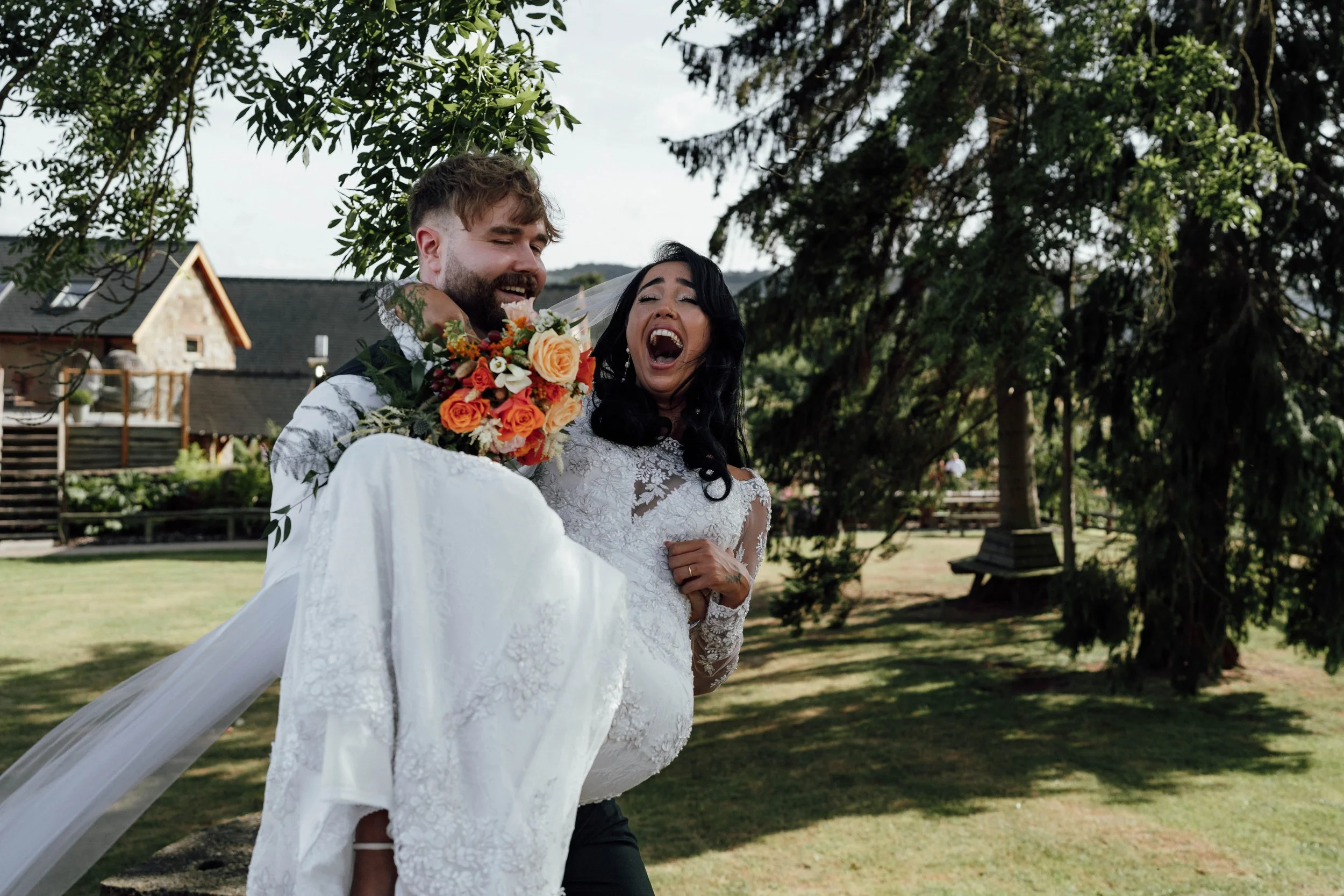A bride and groom celebrating outdoors, with the groom carrying the bride and holding a bouquet of flowers, under trees in a park or garden setting.