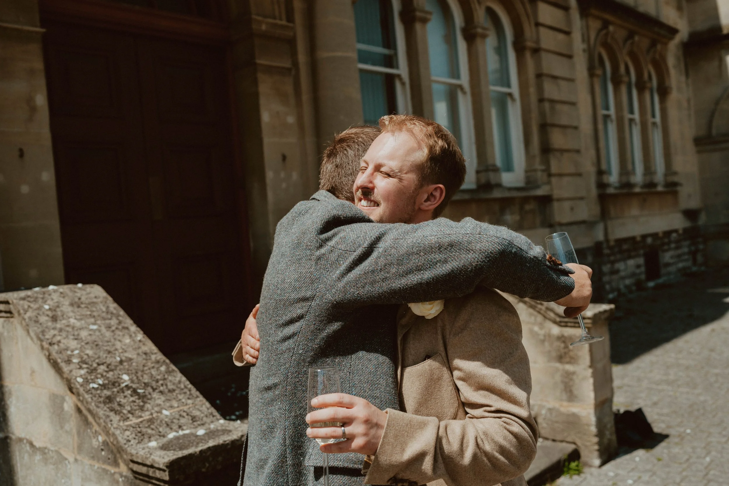 Two men hugging outdoors, one smiling with eyes closed holding a glass of champagne, in front of a historic building.