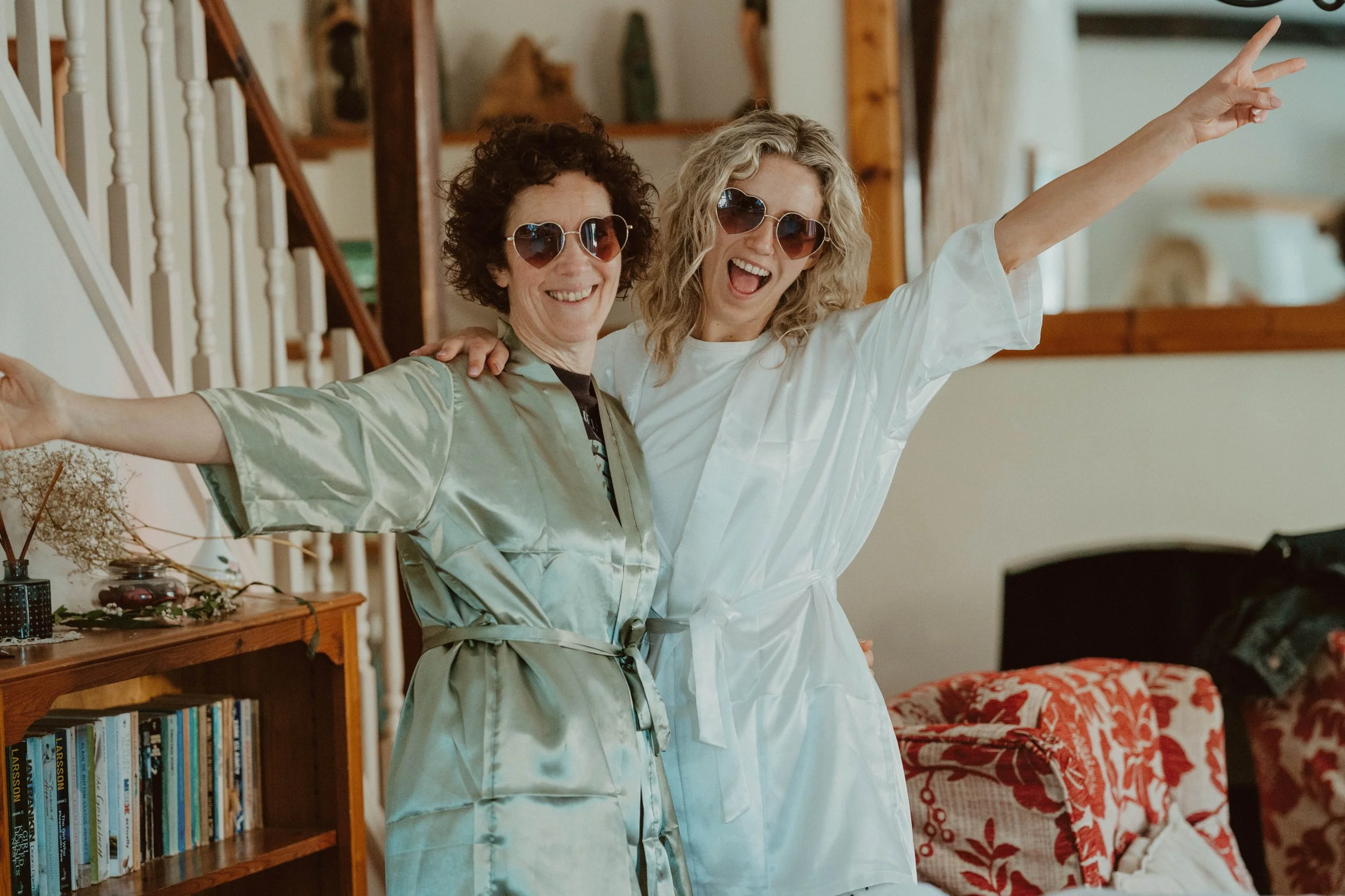 Two women wearing satin robes and heart-shaped sunglasses smiling and celebrating indoors.