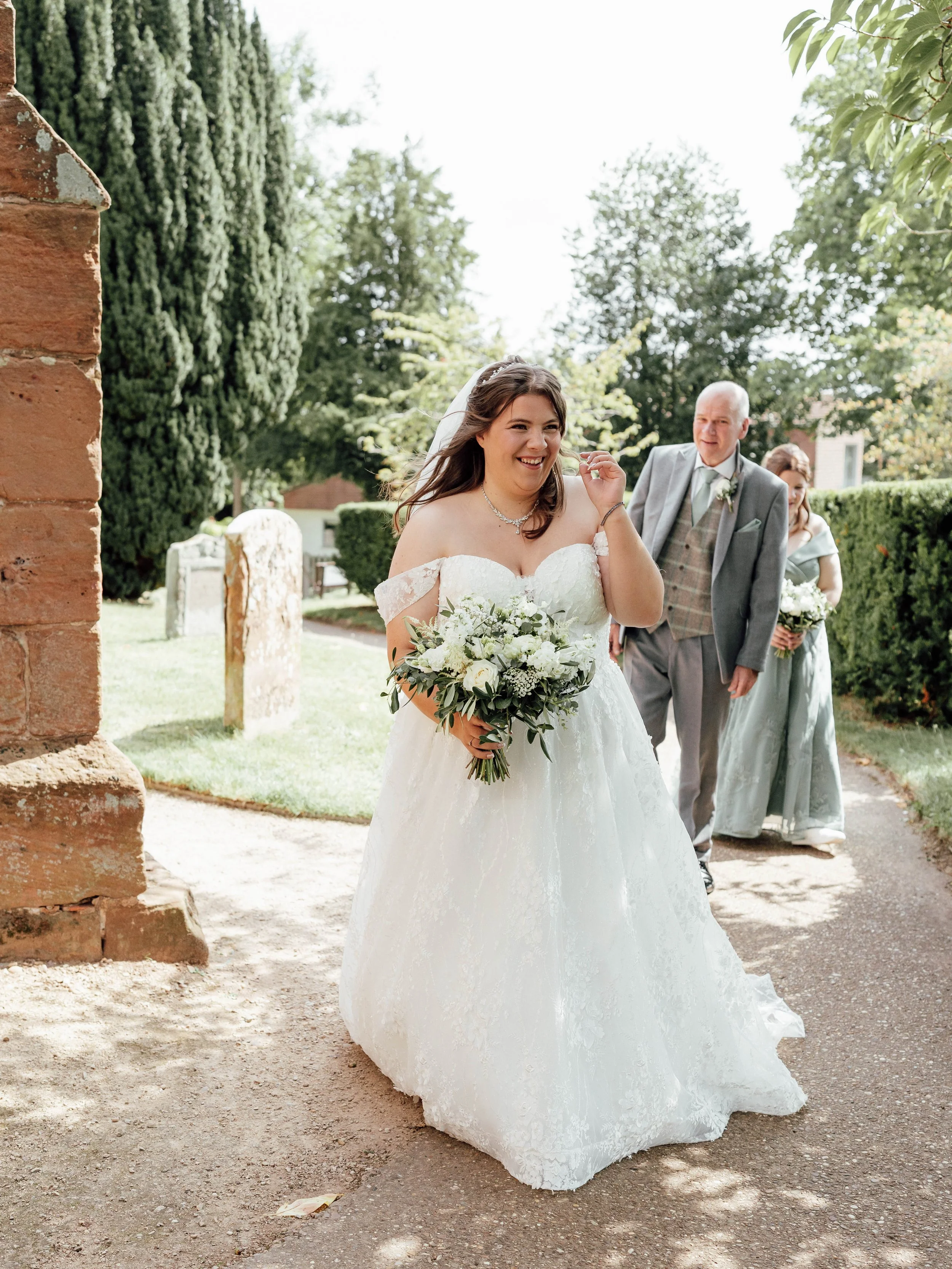 A bride in a white wedding dress holding a bouquet of white and green flowers smiling while walking outdoors on a sunny day, with a man and a woman behind her also carrying bouquets.