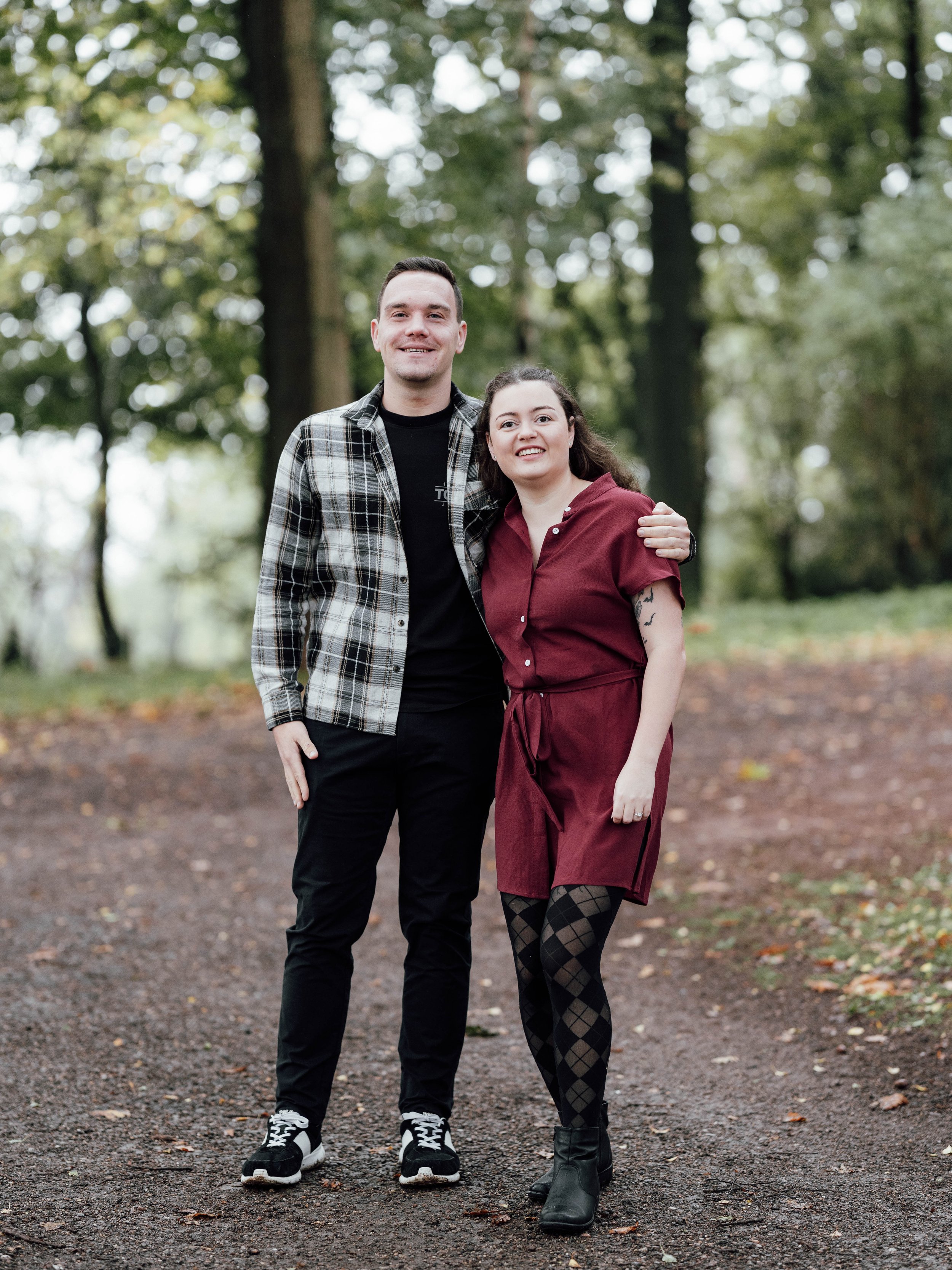 A man and woman standing close together outdoors on a wooded trail, smiling at the camera. The man is wearing a plaid shirt over a black t-shirt, black pants, and sneakers. The woman is wearing a maroon dress, patterned tights, and ankle boots, with 