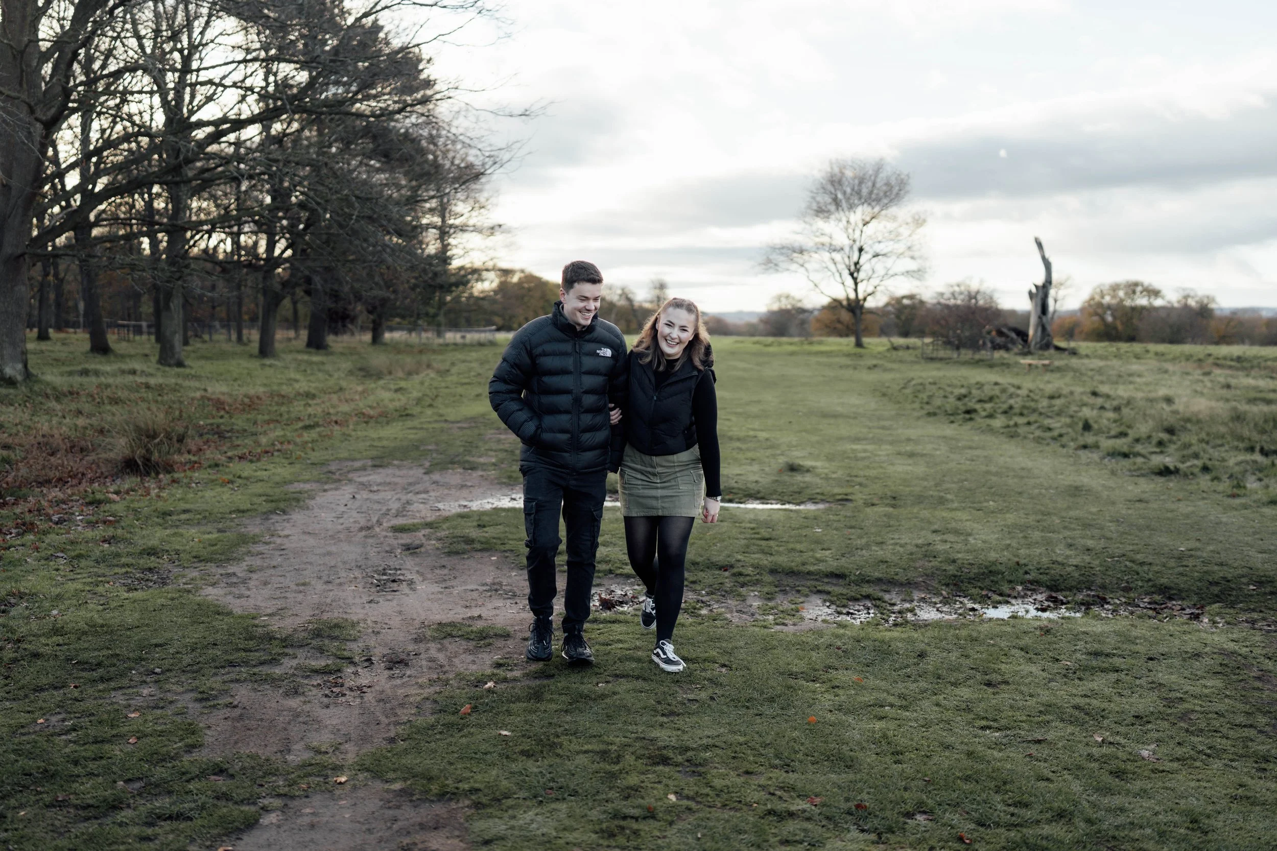 A young man and woman walking arm in arm on a muddy dirt path in a park with trees and cloudy sky in the background.
