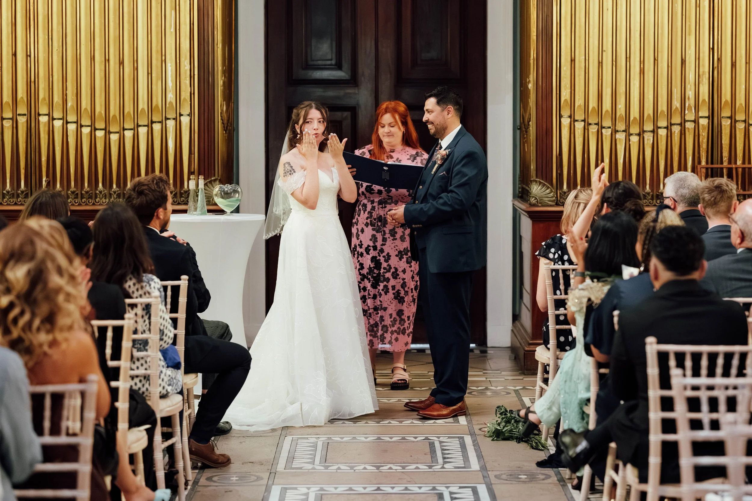 A couple getting married during a wedding ceremony in a decorated indoor venue, with guests seated on both sides of the aisle and a woman officiating.