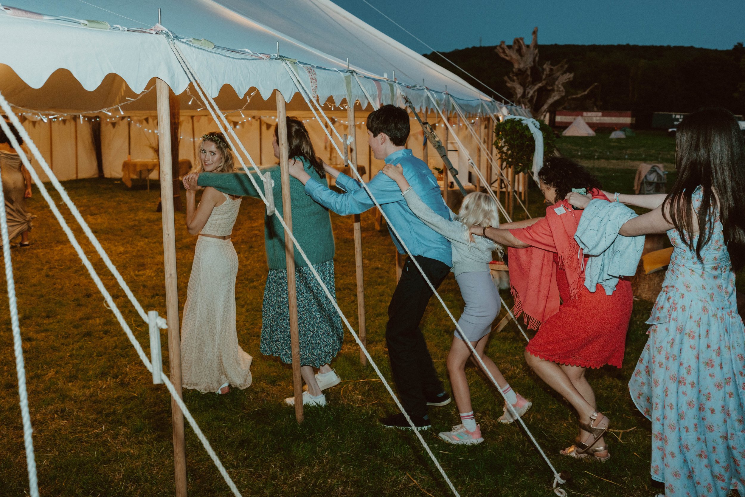 A group of people playing a tug of war game outside during the evening. They are pulling on a rope, with a large tent in the background and a grassy field.