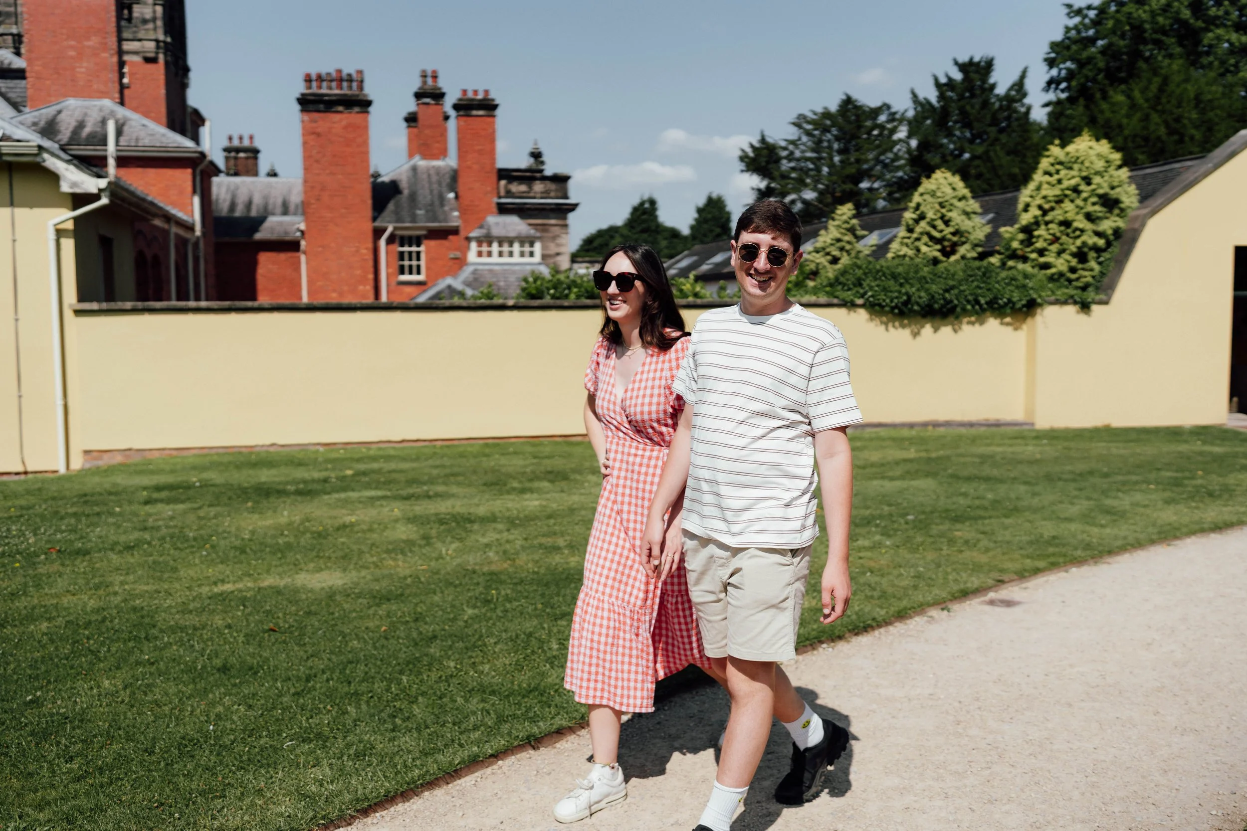 A young man and woman walking outdoors on a sunny day, smiling and wearing sunglasses. The woman is in a red and white checkered dress, and the man is in a white striped t-shirt and shorts. Behind them is a grassy area, a yellow wall, and houses with