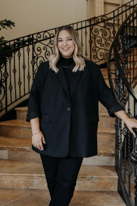 A woman in a black blazer and matching black pants standing on a staircase with a black wrought iron railing, smiling at the camera.