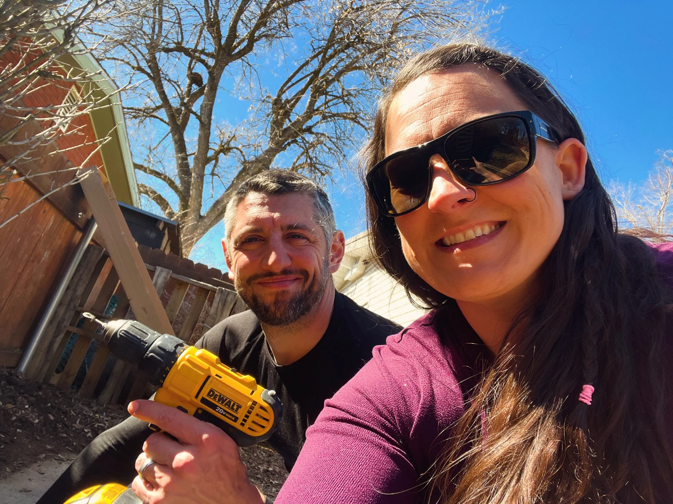 Two people smiling outside in a backyard, one woman with long hair and sunglasses, and a man holding a yellow power drill. There are trees, a wooden fence, and a house in the background.