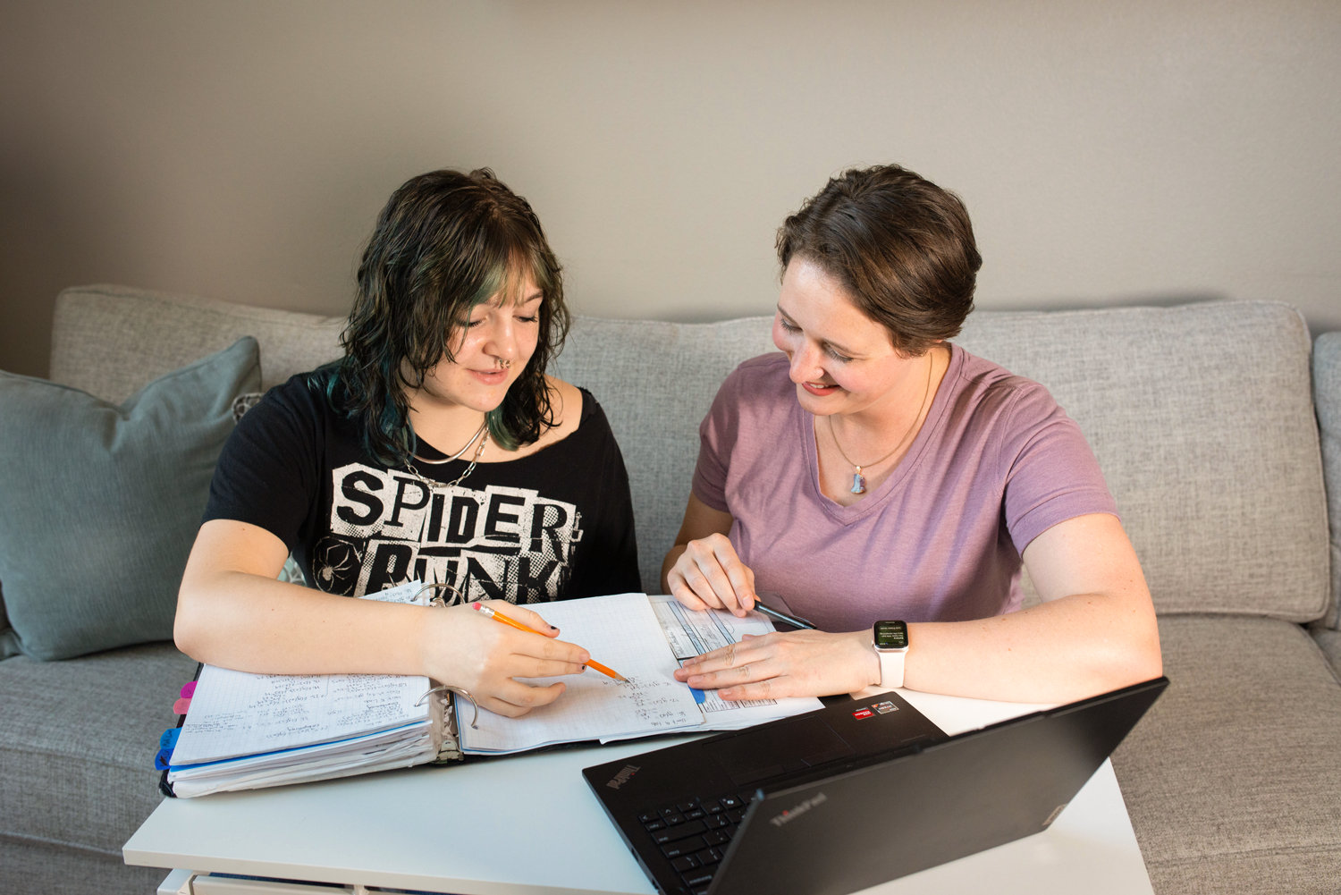 Two women sitting at a table with notebooks, a laptop, and papers, studying together in a living room.