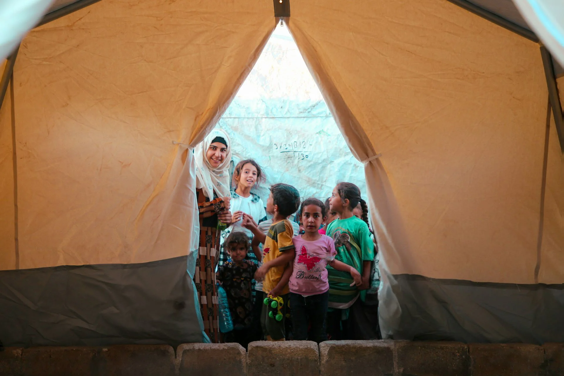 Group of children and a woman inside a tan-colored tent, looking out and smiling at the camera.