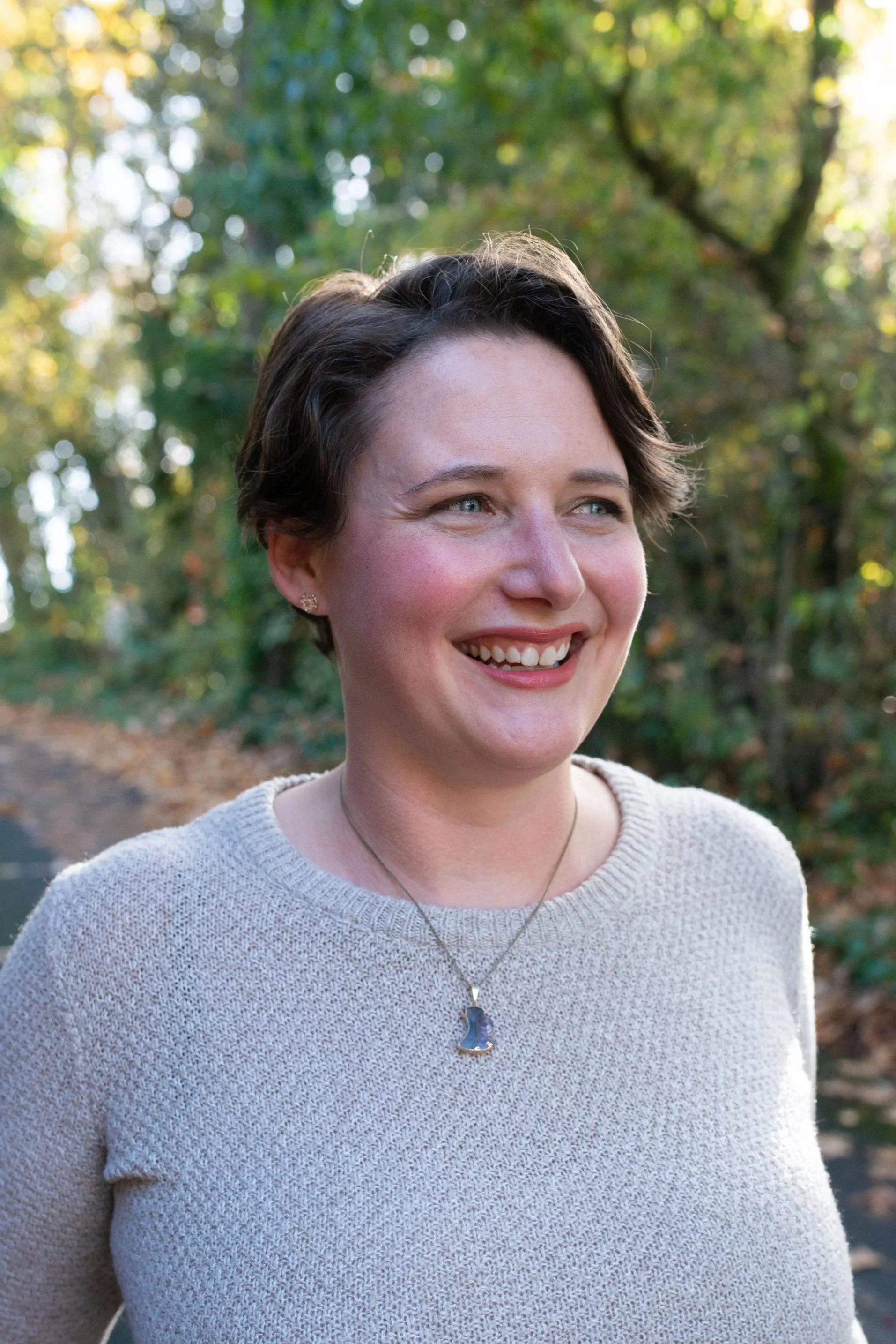 A smiling woman with short dark hair outdoors with blurred trees and sunlight in the background.