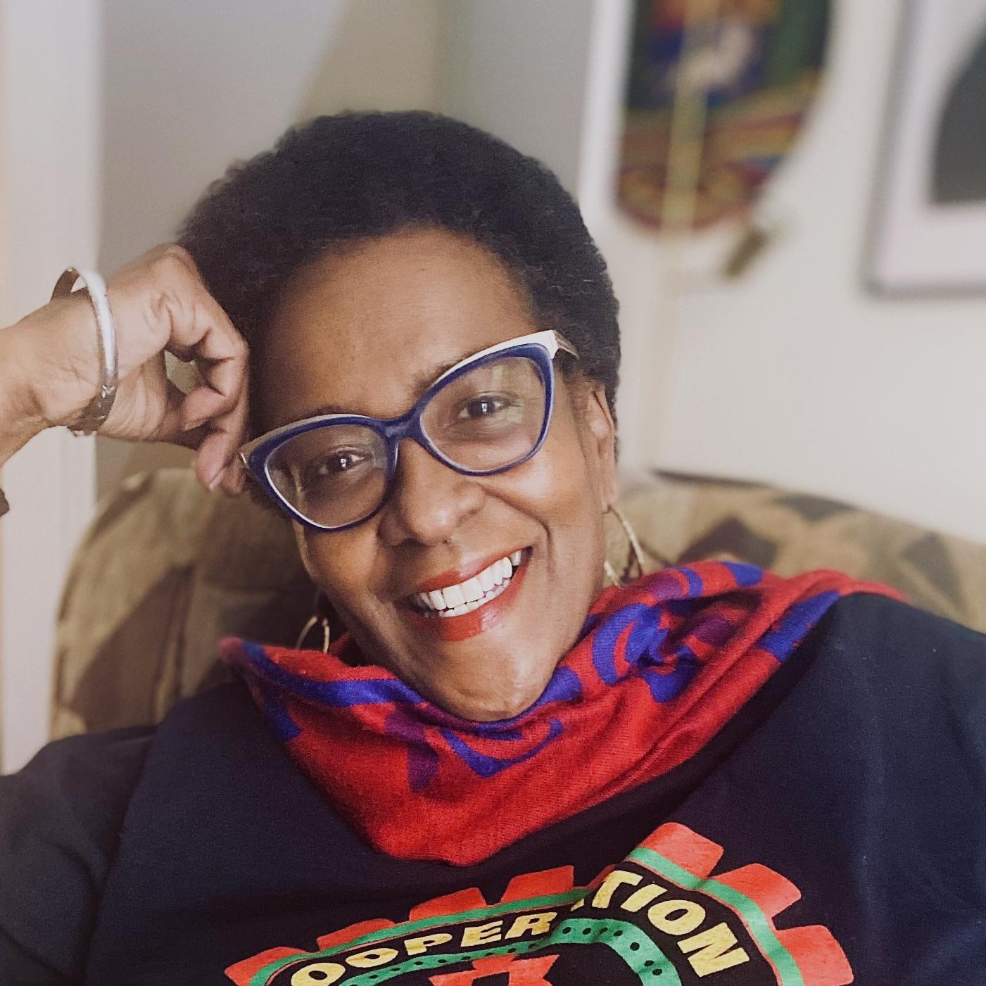 Itah Sadu smiles in an informal portrait with bright red and blue scarf and cats-eye glasses